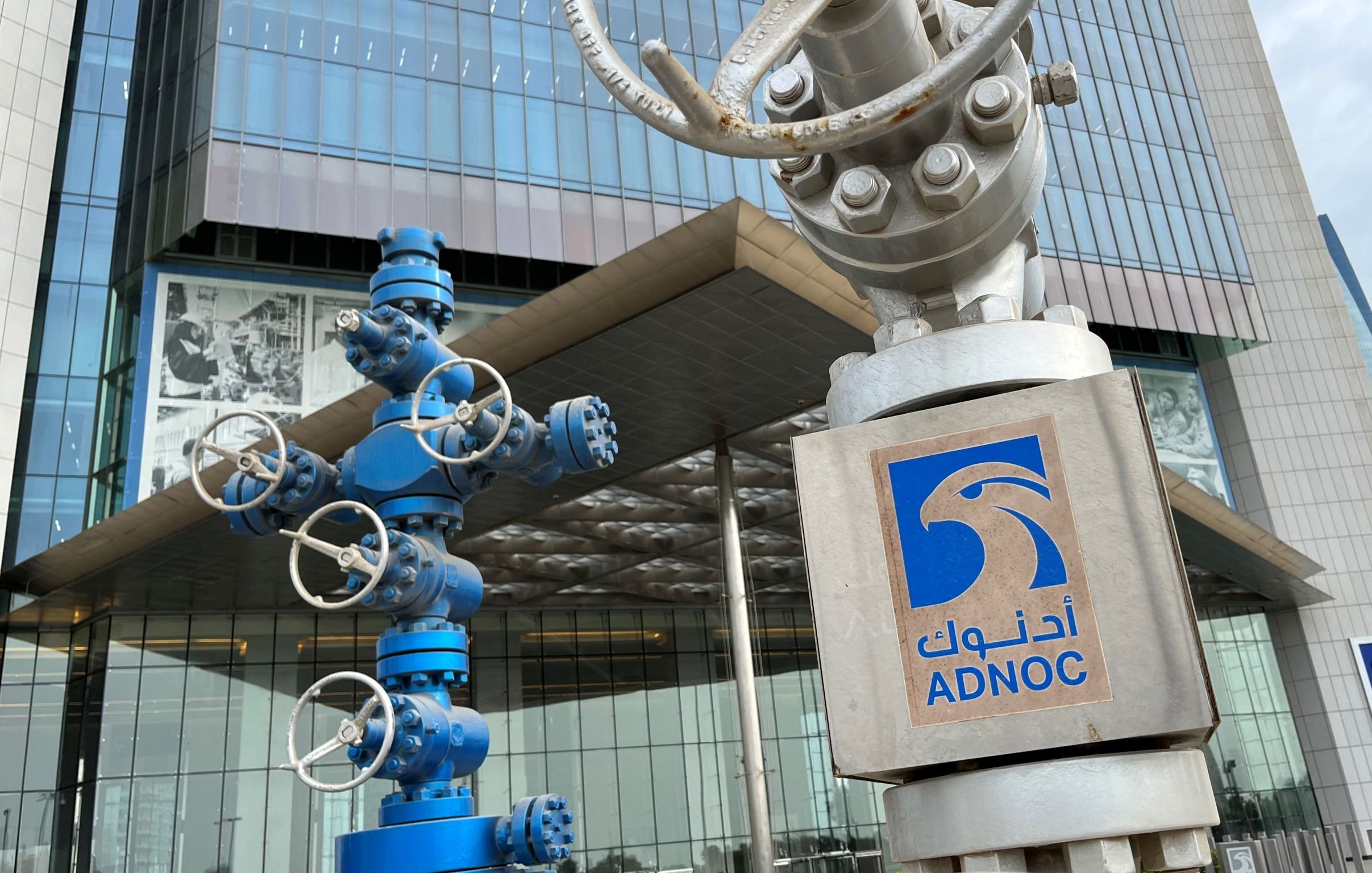 L'image montre un bâtiment moderne avec une façade en verre. À l'avant, on distingue deux équipements de contrôle, généralement utilisés dans le secteur pétrolier. L'un des équipements est de couleur bleue, tandis que l'autre est gris et porte un logo au sommet indiquant "ADNOC", qui est l'acronyme de la Compagnie Pétrolière Nationale des Émirats Arabes Unis. On peut également voir des panneaux avec le nom ADNOC en arabe et en anglais.