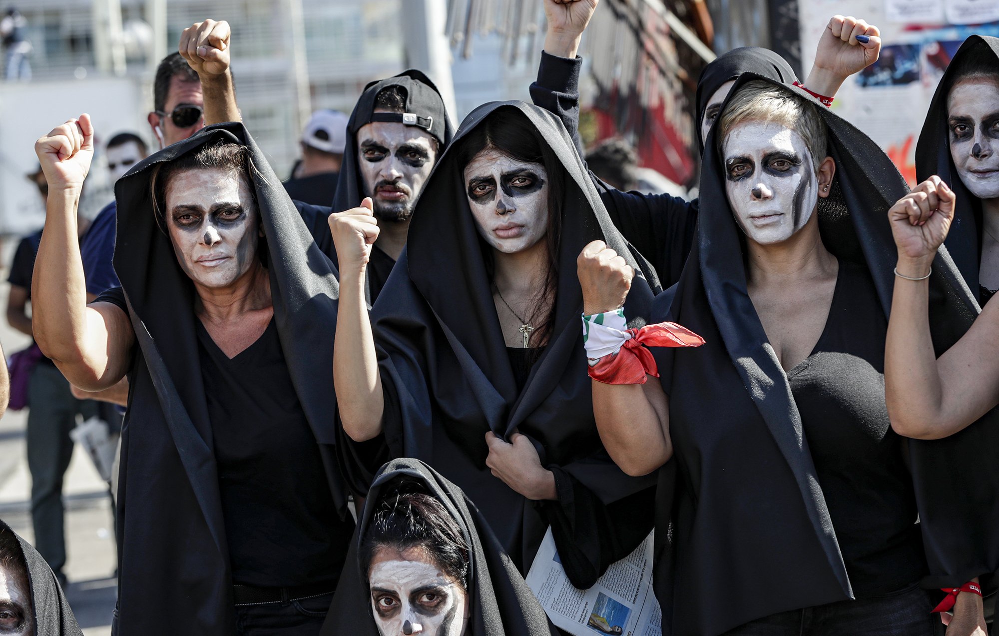 در این تصویر، گروهی از افراد زن و مرد را مشاهده می‌کنیم که لباس‌های سیاهی به تن دارند و صورت‌های خود را با رنگ سفید نقاشی کرده‌اند. آن‌ها در حال نمایش اعتراض خود هستند و برخی از آن‌ها با دستان بلند شده، نشانه‌ای از قدرت و اتحاد را نشان می‌دهند. پس‌زمینه تصویر به وضوح حاکی از یک تجمع یا تظاهرات است. حالت چهره‌ها جدی و معنادار به نظر می‌رسد، که بیانگر عزم و اراده آن‌ها برای بیان دیدگاه‌ها و خواسته‌هایشان است.