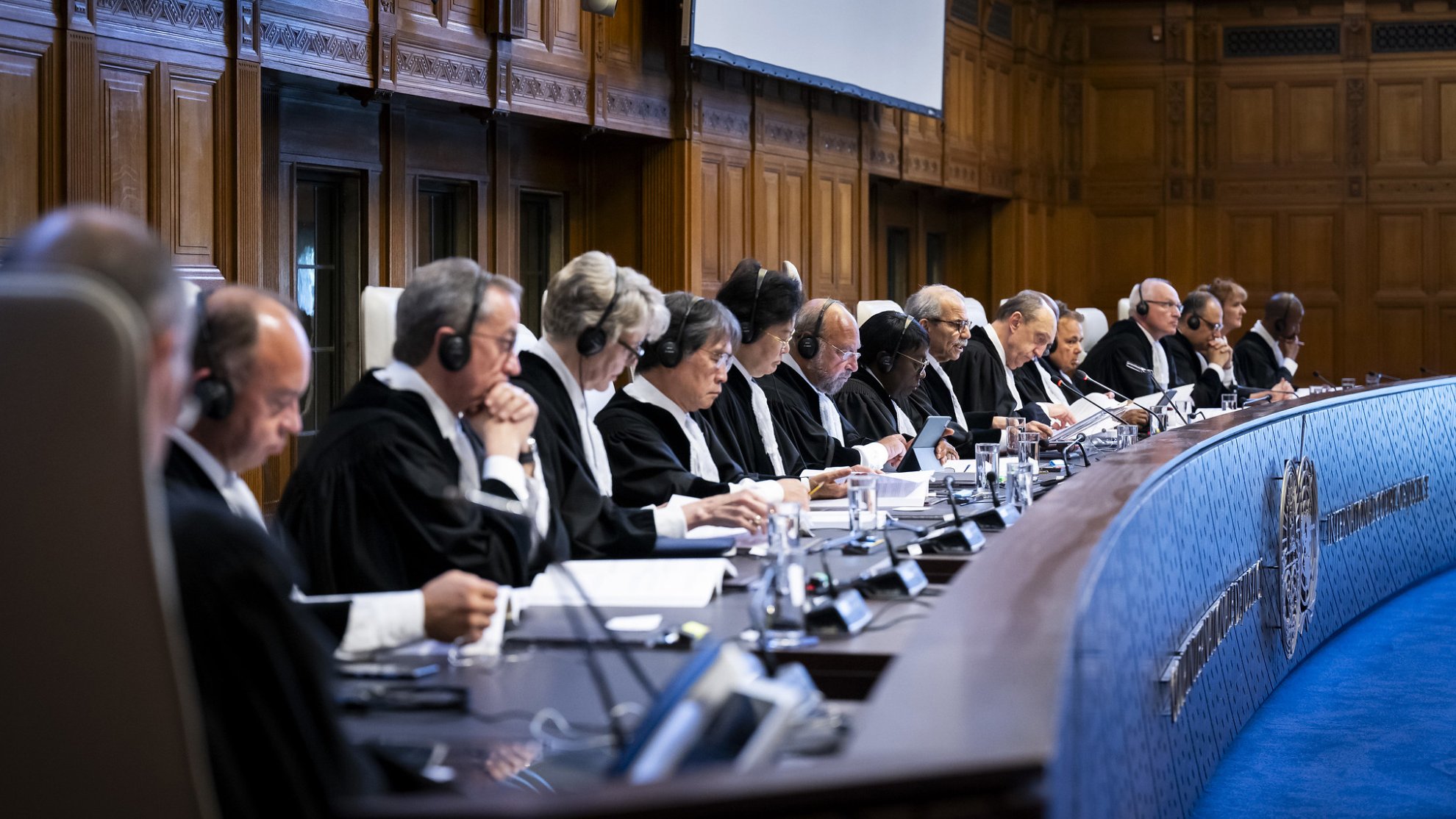 L'image montre une salle de tribunal où plusieurs juges sont assis en ligne dans un espace formel et bien éclairé. Ils portent des robes noires et des écouteurs, symbolisant une séance judiciaire importante. Le décor est élégant, avec des murs en bois et un grand écran au fond. Les juges semblent concentrés sur les documents devant eux, participant à une discussion ou à une audience. L'atmosphère est sérieuse, en accord avec la nature de leurs fonctions.