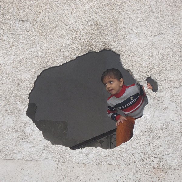 L'image montre un enfant qui regarde à travers un trou dans un mur. Le mur est en mauvais état, avec une surface rugueuse et des fissures visibles. L'enfant porte un pull rayé et semble curieux, se penchant légèrement pour observer ce qui se trouve de l'autre côté du mur. La scène évoque une atmosphère d'exploration et d'intrigue.