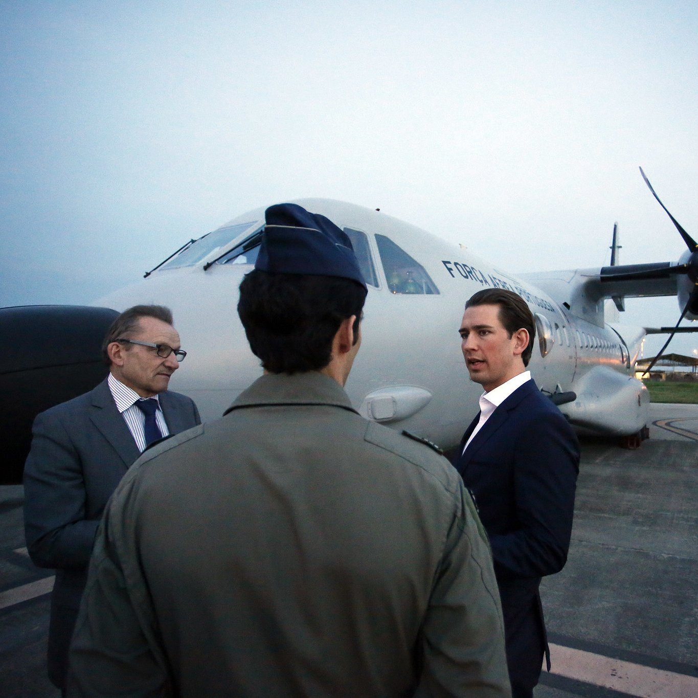 L'image montre trois hommes près d'un avion. L'un des hommes porte un uniforme militaire, tandis que les deux autres sont habillés dans des costumes. Ils semblent être en conversation, face à l'avion de transport, qui est stationné sur le tarmac à la tombée de la nuit. L'éclairage ambiant est doux, et on peut voir des lumières à l'arrière-plan, probablement d'un aéroport ou d'une base aérienne.
