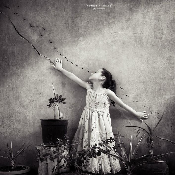 L'image représente une petite fille dans une robe blanche, se tenant debout devant un mur texturé. Elle est en train de tendre les bras, comme si elle essayait d'attraper quelque chose au-dessus d'elle. On peut voir une série d'oiseaux en vol dessinée sur le mur, créant un effet de mouvement. À ses pieds, il y a des plantes en pot qui ajoutent une touche naturelle à la scène. La photographie est en noir et blanc, ce qui lui donne une atmosphère à la fois rêveuse et nostalgique.
