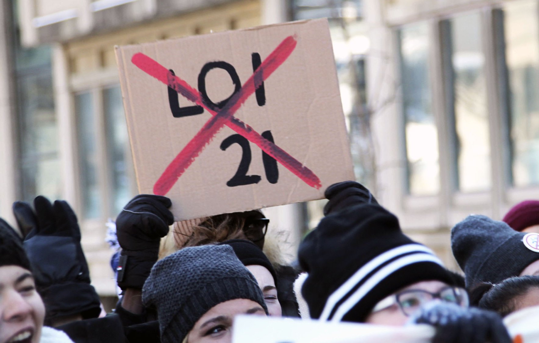 L'image montre un groupe de personnes manifestant. Au premier plan, une personne tient une pancarte en carton sur laquelle est écrit "LOI 21" avec une grande croix rouge qui la barre. Les participants portent des gants et des bonnets, suggérant qu'ils manifestent par temps frais. L'ambiance semble être celle d'une protestation, probablement contre la loi mentionnée.