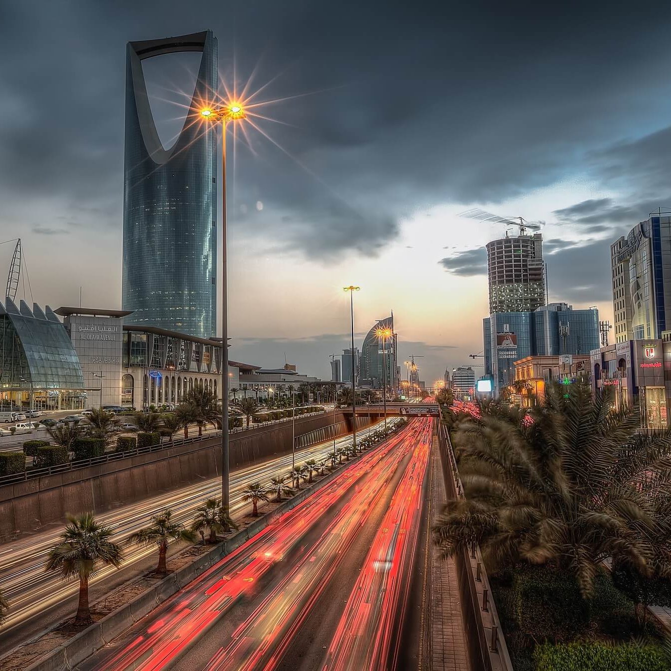 L'image montre une vue urbaine d'une grande ville, probablement Riyad, avec des gratte-ciel modernes qui se dressent contre un ciel nuageux. On peut voir une route animée avec des phares de voitures qui créent des traînées lumineuses, suggérant une circulation intense. Des palmiers sont visibles le long de la route, ajoutant une touche naturelle à ce paysage urbain. La scène est éclairée par des lampadaires, donnant une atmosphère à la fois dynamique et contemporaine.