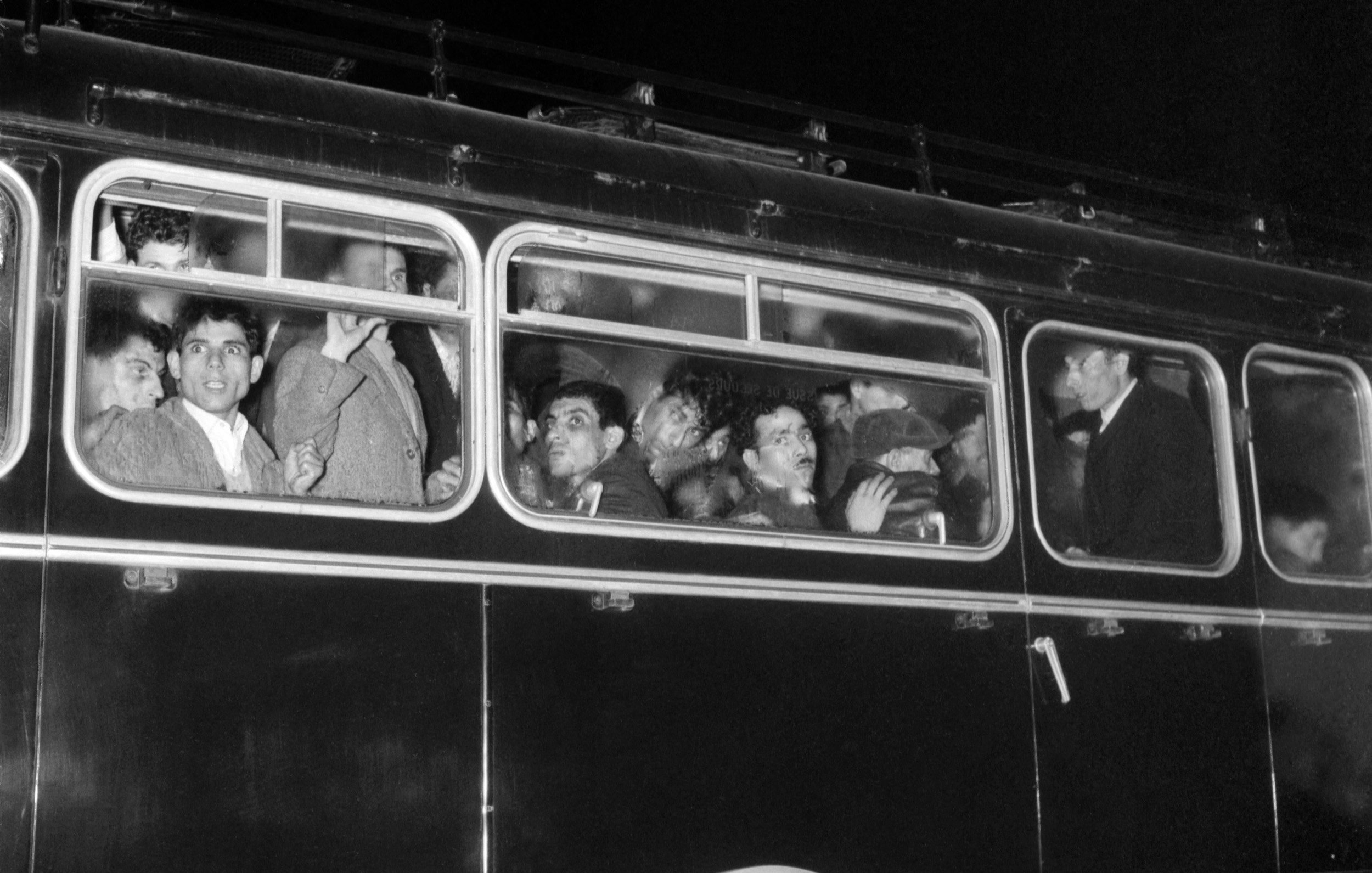 L'image montre un bus noir en soirée, dont les fenêtres sont remplies de passagers. On peut voir plusieurs visages, certains semblent surpris ou curieux, tandis que d'autres affichent des expressions neutres. L'éclairage est faible, accentuant l'atmosphère nocturne de la scène. Le bus a l'air ancien, avec une carrosserie en métal et des détails rétro.