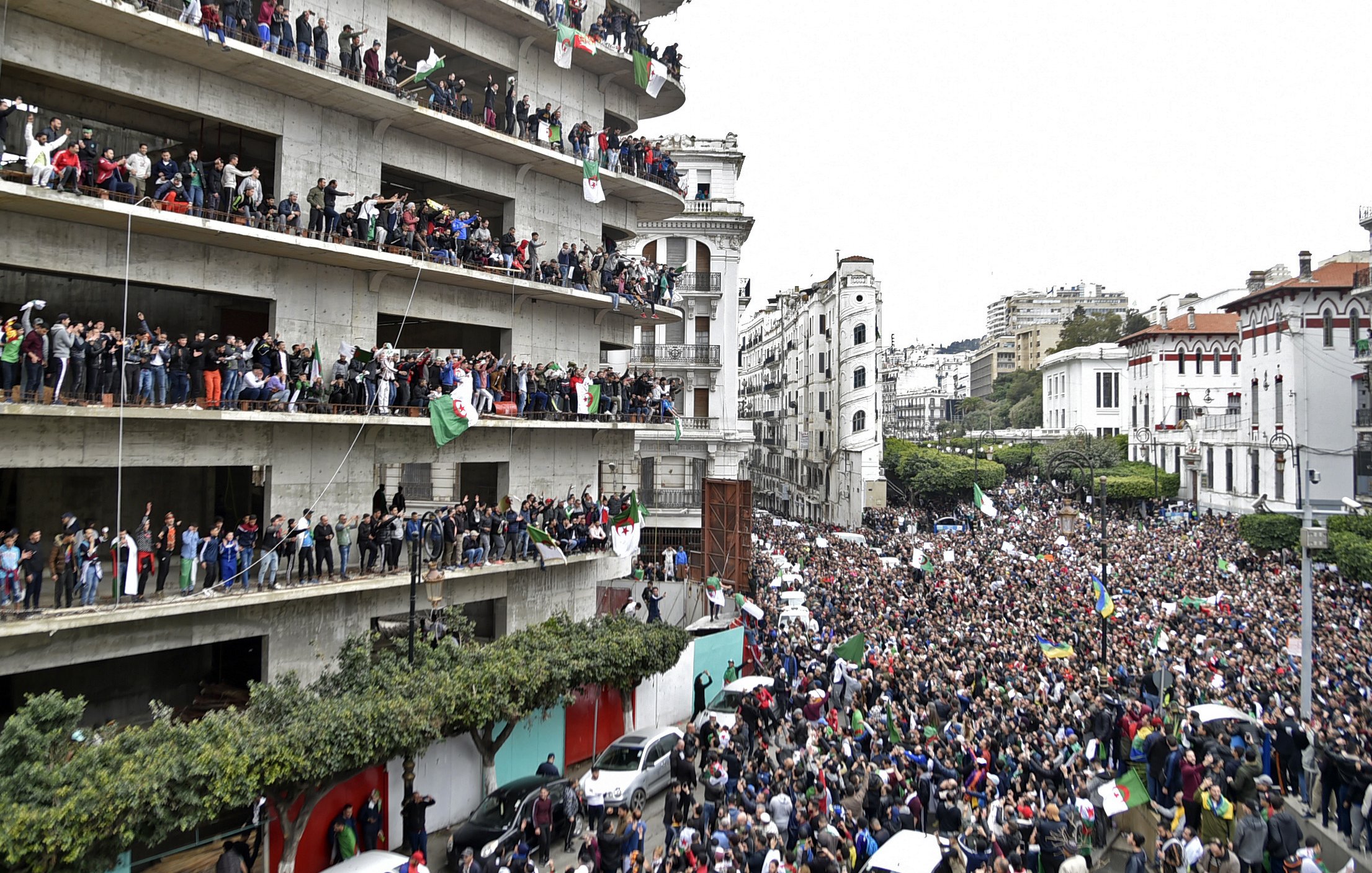 L'image montre une grande foule rassemblée devant un bâtiment, manifestant dans une ville. De nombreuses personnes sont visibles sur les balcons du bâtiment, brandissant des drapeaux et exprimant leur soutien à la manifestation. La rue est remplie de manifestants, et l'atmosphère semble vibrante et dynamique, avec un mélange de drapeaux et de pancartes. Le paysage urbain environnant, composé de divers bâtiments, ajoute à l'environnement de la scène.