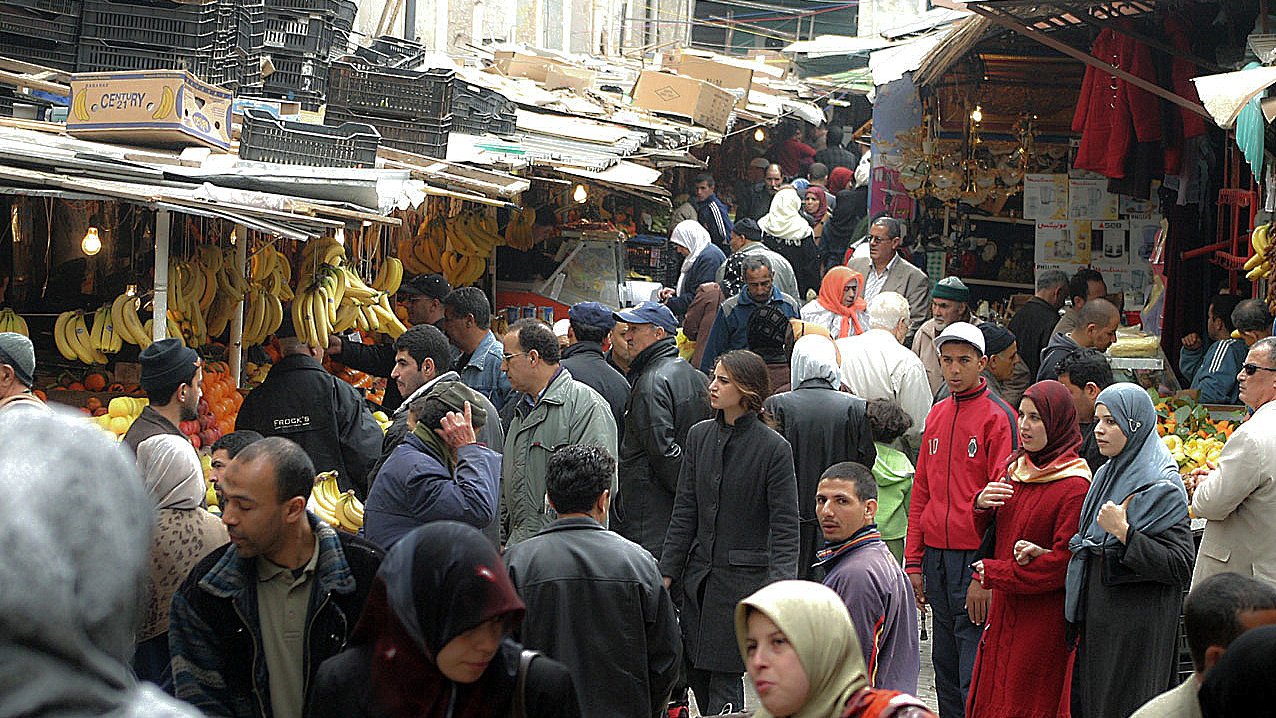 L'image montre un marché animé, probablement dans une ville du Moyen-Orient ou d'Afrique du Nord. Des stands sont remplis de fruits, notamment des bananes et des oranges. On peut voir de nombreuses personnes, vêtues de vêtements variés, se déplacer entre les étals. L'atmosphère est animée, avec des groupes discutant et des clients choisissant des marchandises. Des éclairages pendent du plafond, ajoutant une ambiance particulière au lieu.