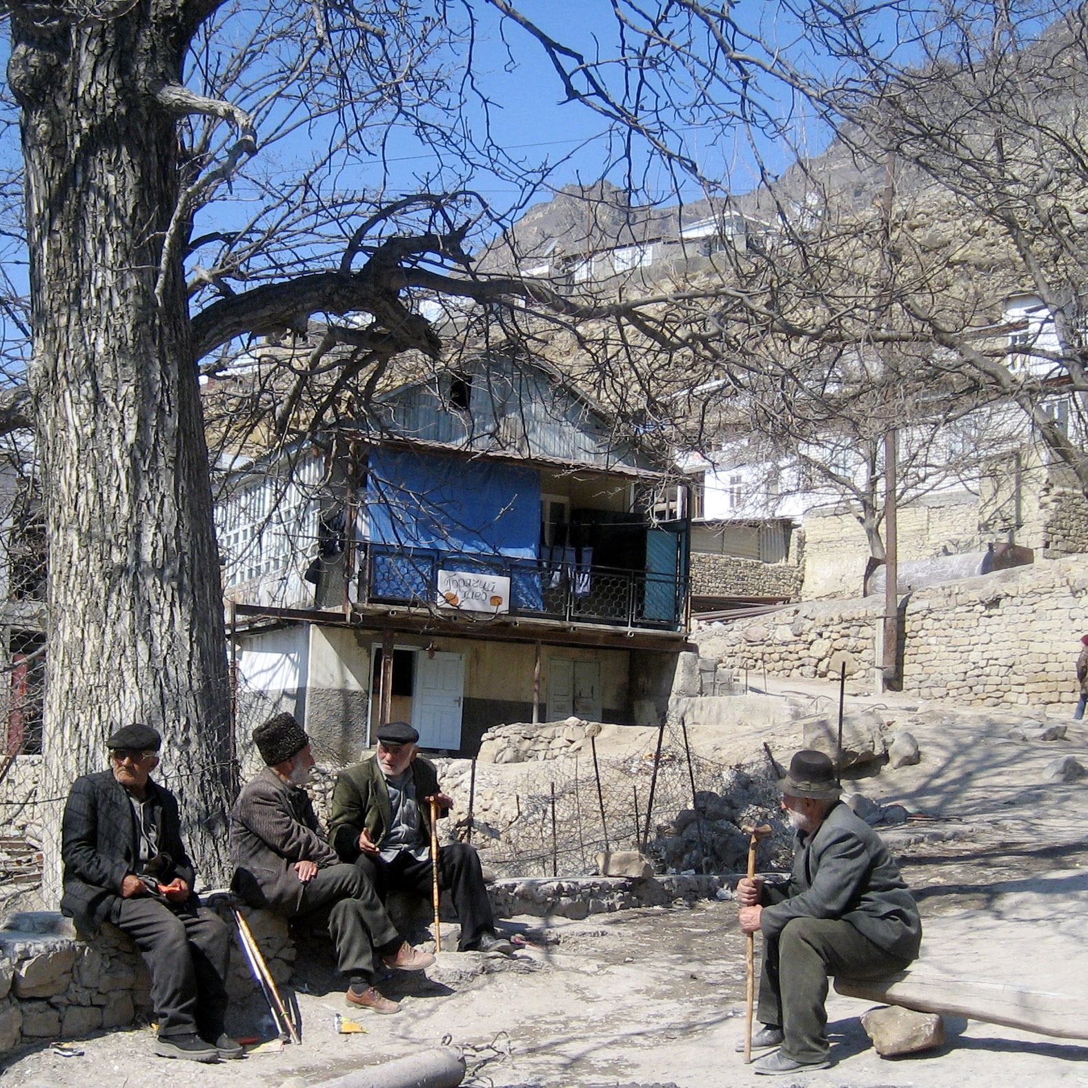 L'image montre un paysage rural avec des maisons en pierre, certaines d'entre elles ayant des balcons. Au premier plan, quatre hommes assis discutent ensemble, habillés de manière traditionnelle. Ils sont entourés d'arbres dénudés, ce qui indique probablement le début du printemps ou l'hiver. L'ambiance semble calme et tranquille, avec un chemin en terre qui serpente entre les maisons.