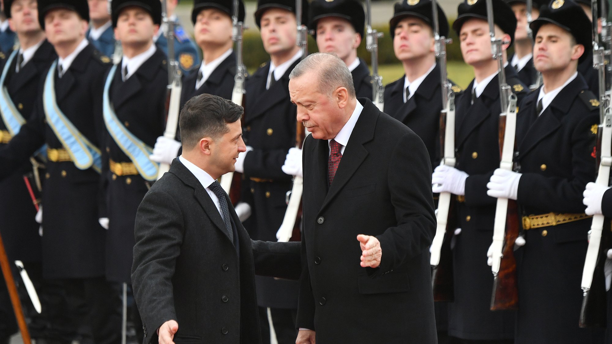 L'image montre deux hommes en discussion. L'un est vêtu d'un manteau noir, tandis que l'autre porte un costume avec une cravate. En arrière-plan, des soldats en uniforme présentent les honneurs, debout et alignés, avec des armes à la main. L'atmosphère semble solennelle, indiquant probablement une rencontre officielle ou un événement diplomatique.