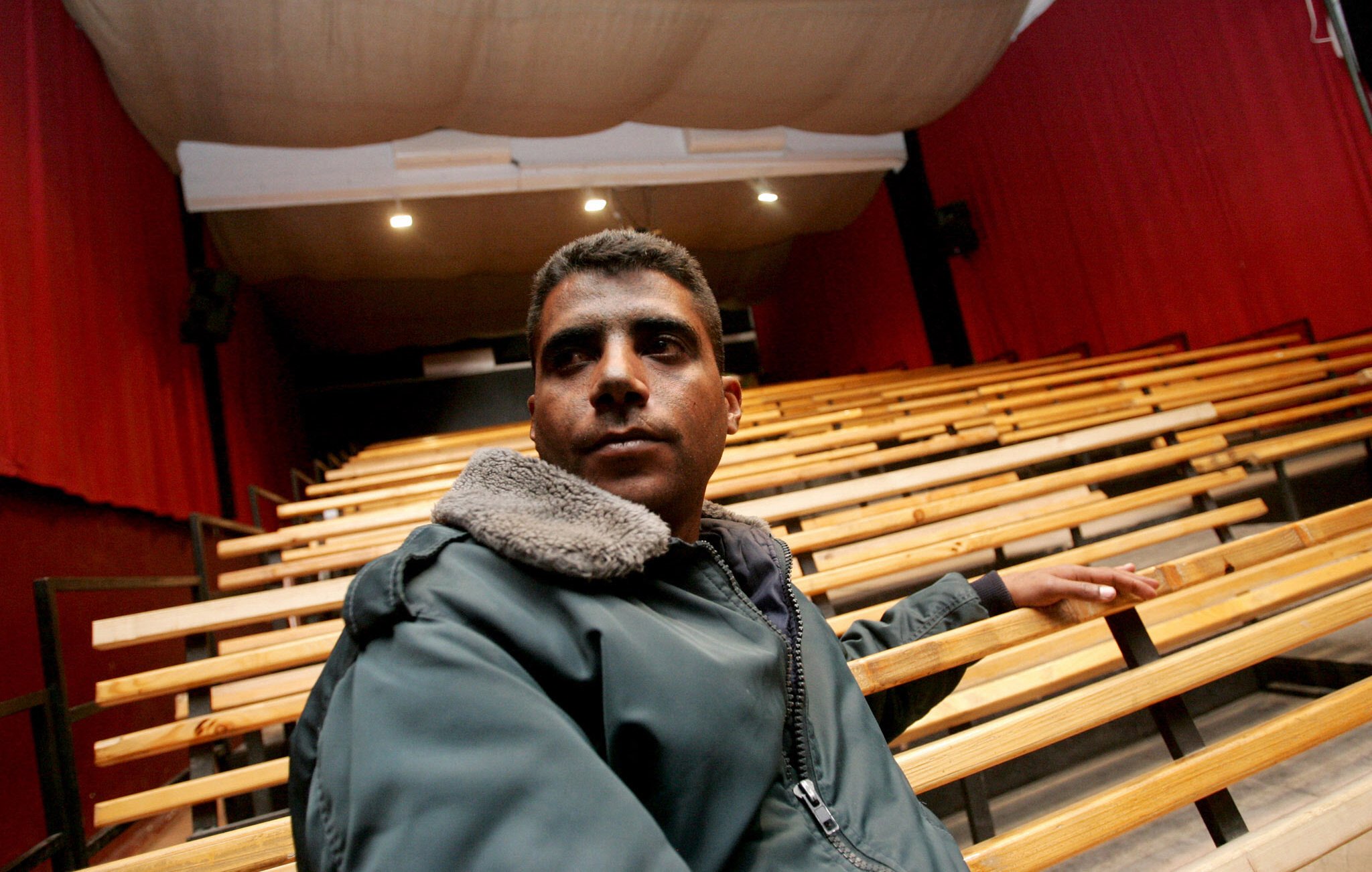 L'immagine mostra un giovane uomo seduto su una panca di legno in un teatro. Il teatro ha un'illuminazione soffusa, con tende rosse che coprono le pareti e un palcoscenico visibile sullo sfondo. L'uomo indossa una giacca scura con un colletto di pelliccia. La sua espressione sembra seria e contemplativa mentre guarda verso l'esterno, evidenziando un'atmosfera di introspezione nel contesto di un luogo culturale. Le file di sedili vuoti suggeriscono un ambiente che sta aspettando un evento o una performance.