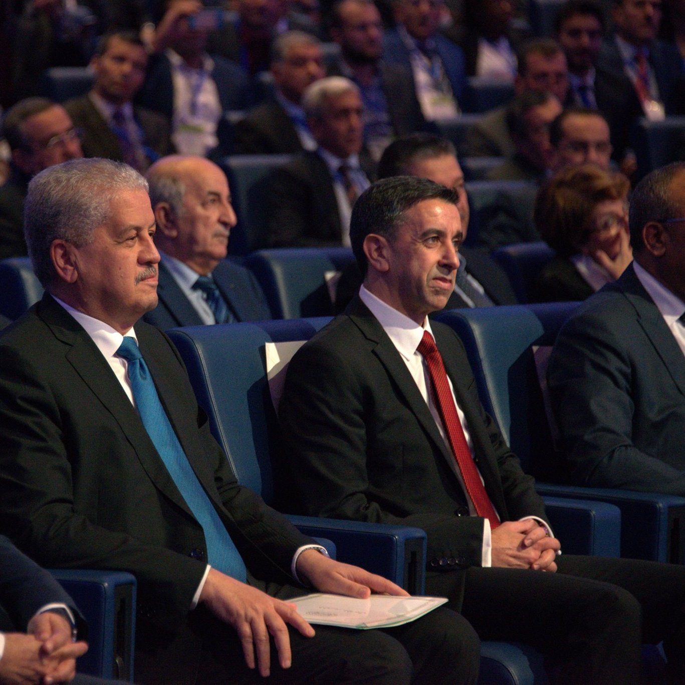 L'image montre un groupe d'hommes assis dans une salle de conférence, apparemment lors d'un événement officiel. Ils portent des costumes sombres, avec une attention particulière sur deux hommes au premier plan, l'un en costume noir avec une cravate bleue, et l'autre en costume noir avec une cravate rouge. En arrière-plan, on peut voir d'autres personnes, probablement des participants ou des invités, attentifs à ce qui se passe sur scène. L'atmosphère semble formelle et sérieuse.