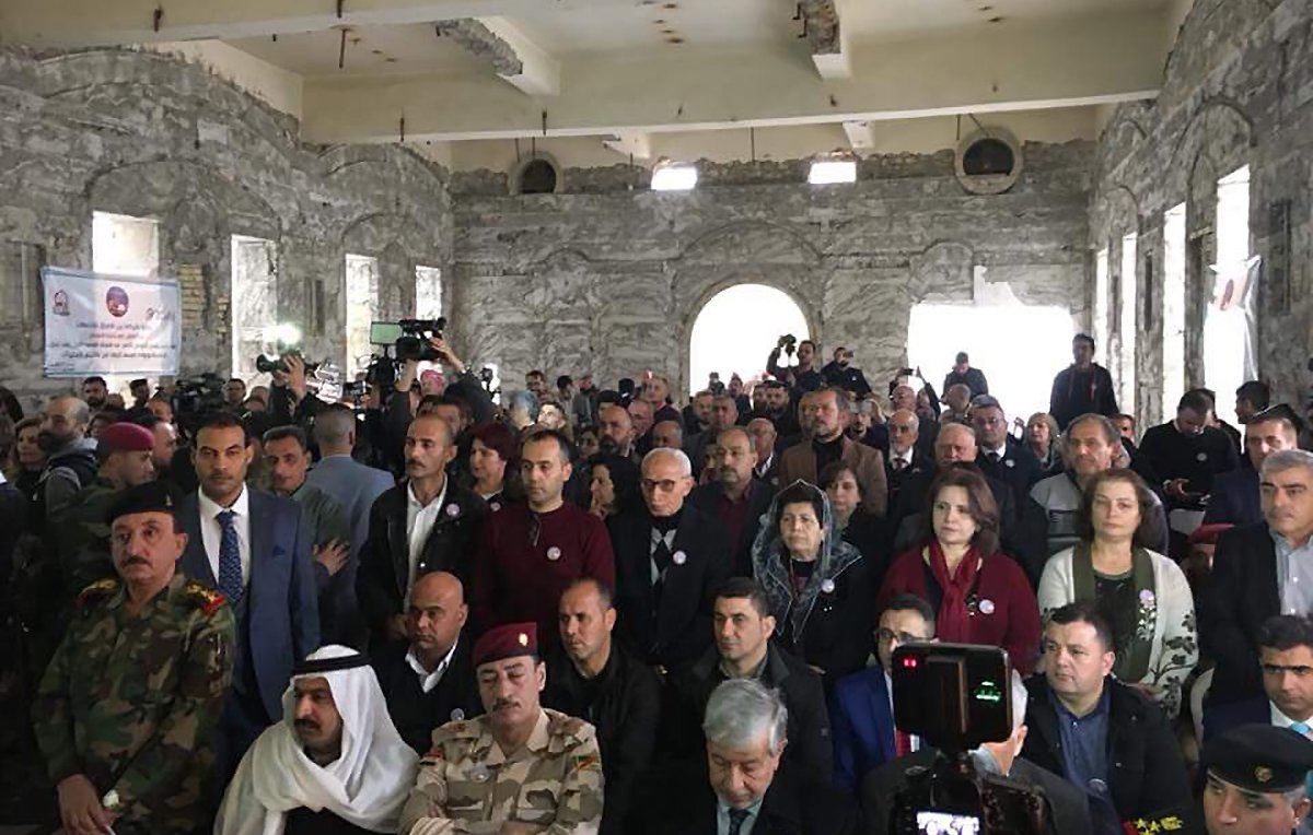 L'image montre une grande foule de personnes rassemblées dans un bâtiment à l'apparence industrielle ou historique, avec des murs en pierre. Les participants semblent être assis ou debout et portent des badges ou des pin's. Diverses personnes de différentes origines apparaissent, y compris des représentants militaires en uniforme. L'atmosphère semble formelle, suggérant un événement significatif ou une réunion. Des caméras et des journalistes sont également présents, indiquant qu'il pourrait s'agir d'une conférence ou d'une célébration.