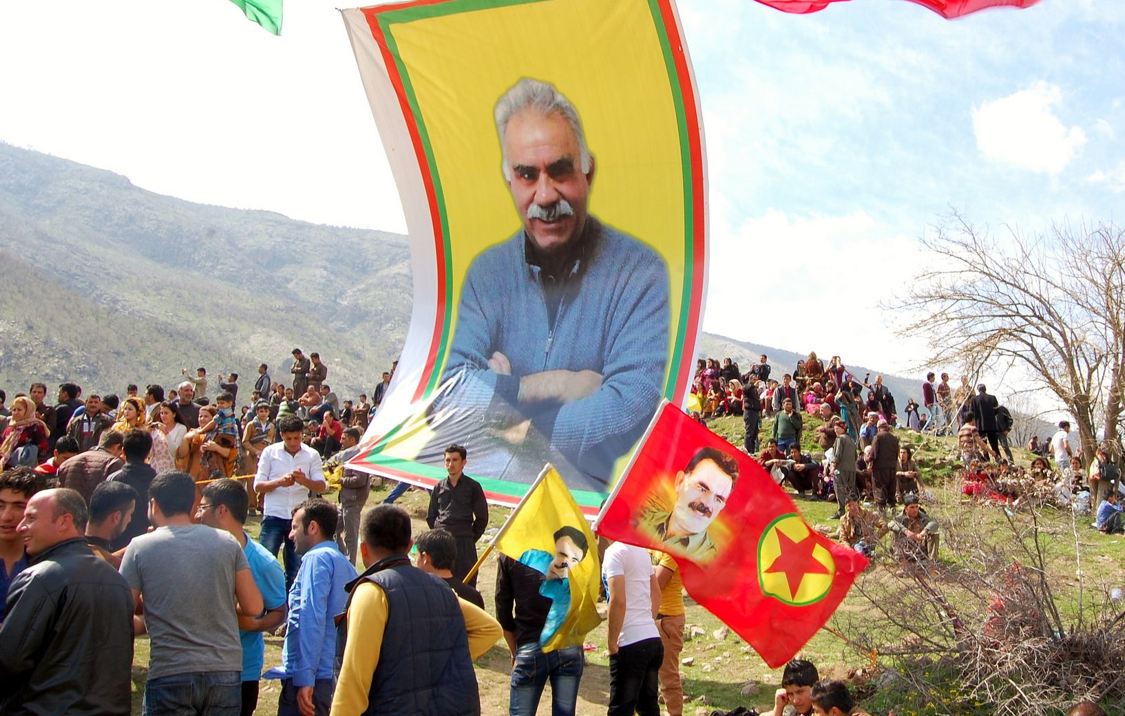 L'image montre une grande réunion en plein air, probablement lors d'une célébration ou d'un événement politique. On peut voir un grand drapeau avec le portrait d'un homme, entouré de plusieurs drapeaux aux couleurs vives, notamment du jaune et du rouge. De nombreuses personnes sont rassemblées et semblent animées, certaines brandissant des drapeaux. En arrière-plan, des montagnes et des arbres se dressent, suggérant un cadre naturel. L'atmosphère semble festive et engagée.