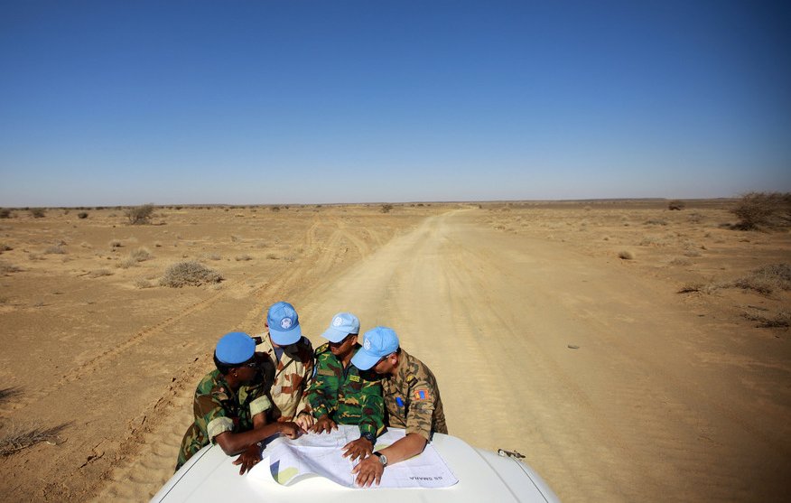 L'image présente un groupe de personnes, probablement des soldats de la paix, qui se penchent sur une carte étalée sur le toit d'un véhicule. Ils portent des casquettes de l'ONU et des uniformes militaires. L'arrière-plan montre un paysage désertique, avec un sol sablonneux et peu de végétation, sous un ciel ensoleillé et dégagé. La scène semble se dérouler dans une région aride, éloignée, où ces individus discutent des informations cartographiques.