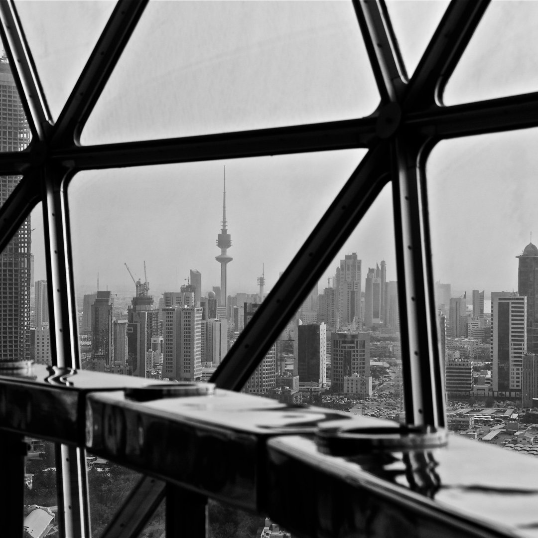 L'image montre une vue urbaine à travers une grande fenêtre en verre avec un cadre géométrique. Au premier plan, on aperçoit une surface réfléchissante et des éléments architecturaux. À l'extérieur, on découvre un paysage de ville en noir et blanc, avec des gratte-ciels modernes et une tour emblématique qui se dresse au loin, probablement un symbole de la skyline. La scène dégage une atmosphère à la fois contemporaine et dynamique.