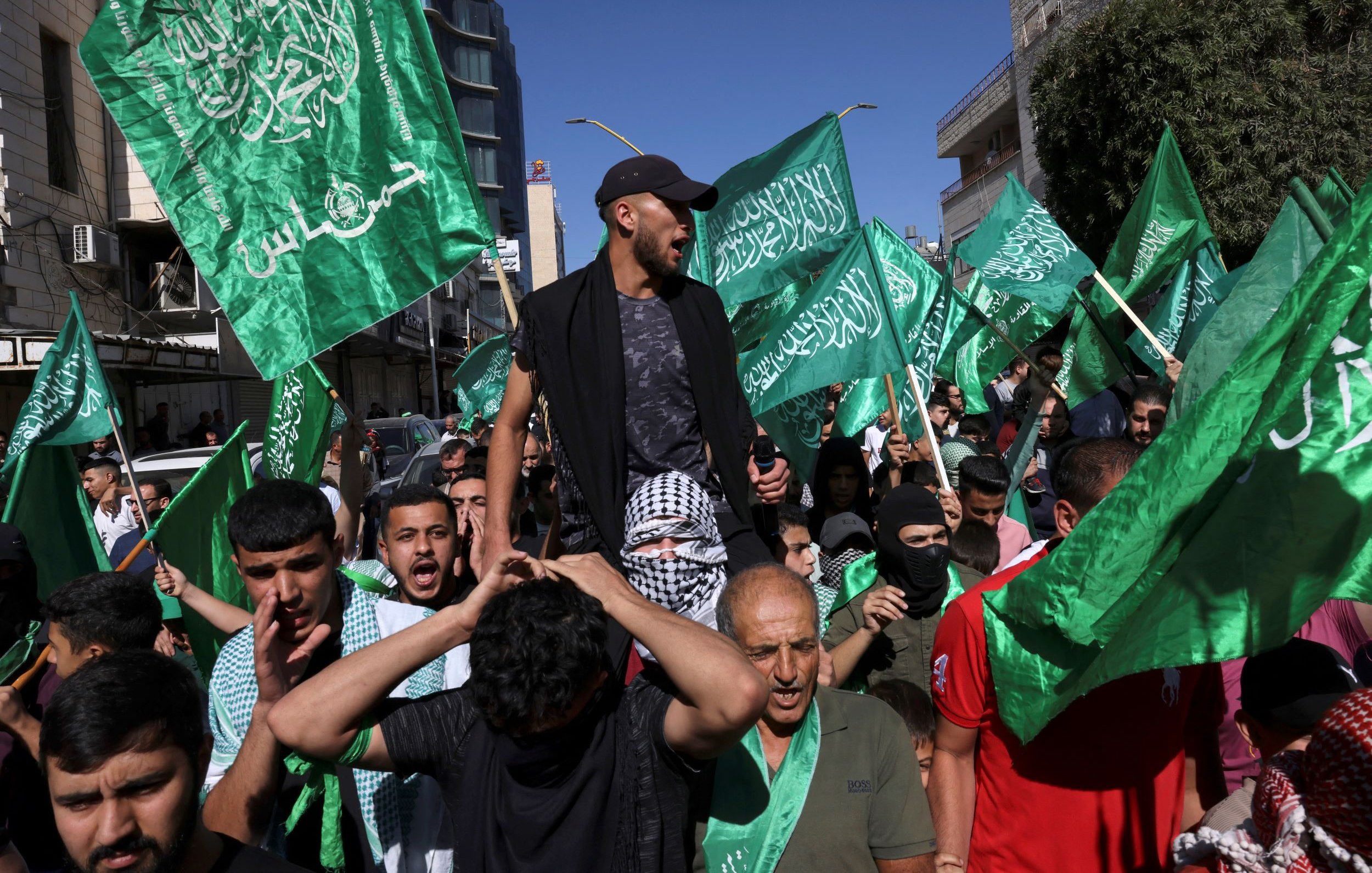 Cette image montre une manifestation animée, avec des personnes brandissant de grands drapeaux verts. On peut voir une foule de manifestants, certains sont en train de crier ou de chanter, tandis qu'un homme est porté sur les épaules de quelqu'un d'autre. L'ambiance semble énergique, et l'arrière-plan présente des bâtiments urbains.