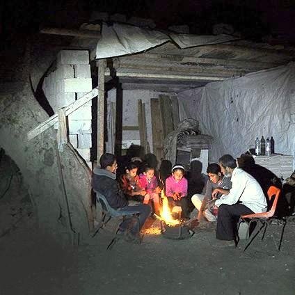 L'image montre un groupe de personnes assises autour d'un feu dans un environnement sombre. Ils semblent rassemblés à l'intérieur d'une structure rudimentaire, faite de matériaux simples comme du bois et des tôles. Les gens, probablement des adultes et des enfants, sont concentrés sur le feu, qui éclaire leur visage et crée une atmosphère chaleureuse malgré les conditions environnantes. Des éléments comme des bouteilles sont visibles en arrière-plan, suggérant une ambiance de partage et de convivialité, malgré le manque de confort.