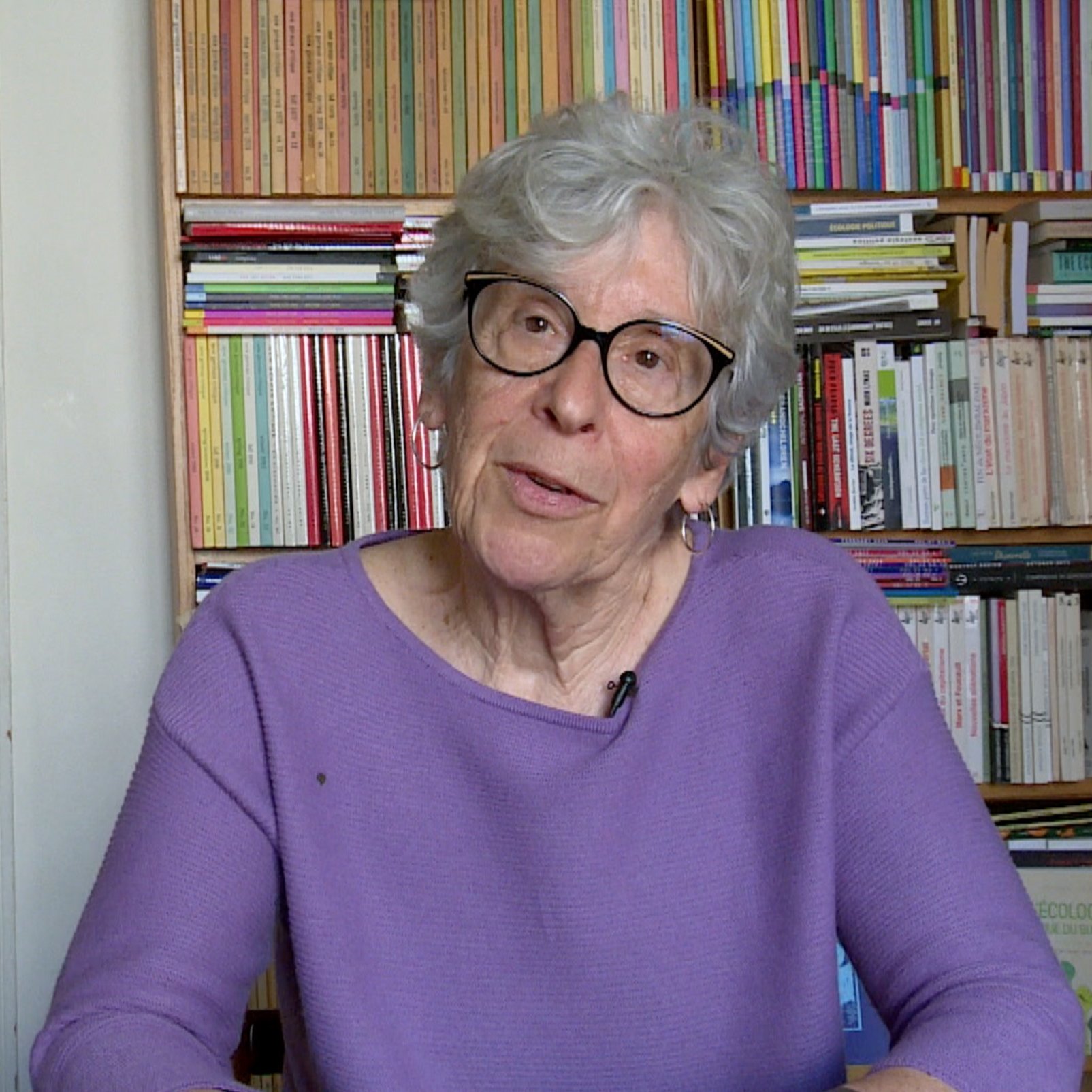 L'image montre une femme âgée avec des cheveux gris et des lunettes. Elle porte un pull violet et est assise devant une bibliothèque remplie de livres, qui présentent des couvertures colorées. Son expression est réfléchie et elle semble engagée dans une conversation. L'environnement est chaleureux et intellectuel, avec des étagères bien ordonnées autour d'elle.