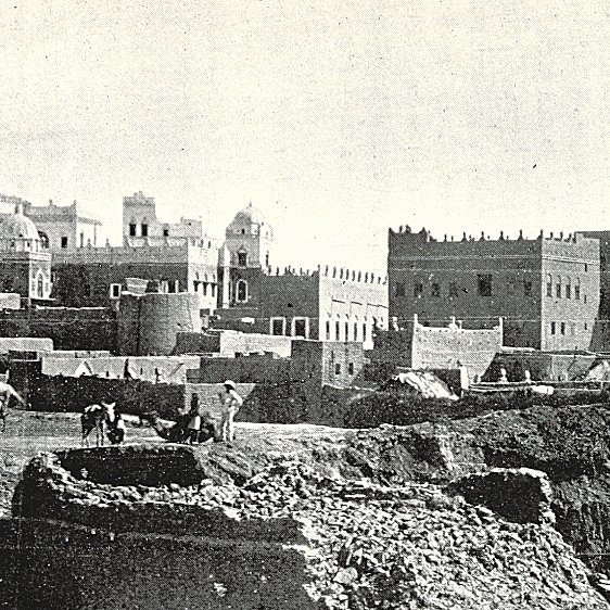 L'image présente un paysage urbain ancien, probablement d'une ville historique. On peut voir des bâtiments en pierre avec des détails architecturaux variés, tels que des tours et des terrasses. À l'avant-plan, on aperçoit des travailleurs, probablement engagés dans des activités agricoles ou de construction. La scène évoque une atmosphère à la fois rustique et animée, avec un ciel clair et des montagnes en arrière-plan qui ajoutent à la dimension historique et géographique de l'endroit. L'ensemble dégage une impression de temps révolu et de tradition.
