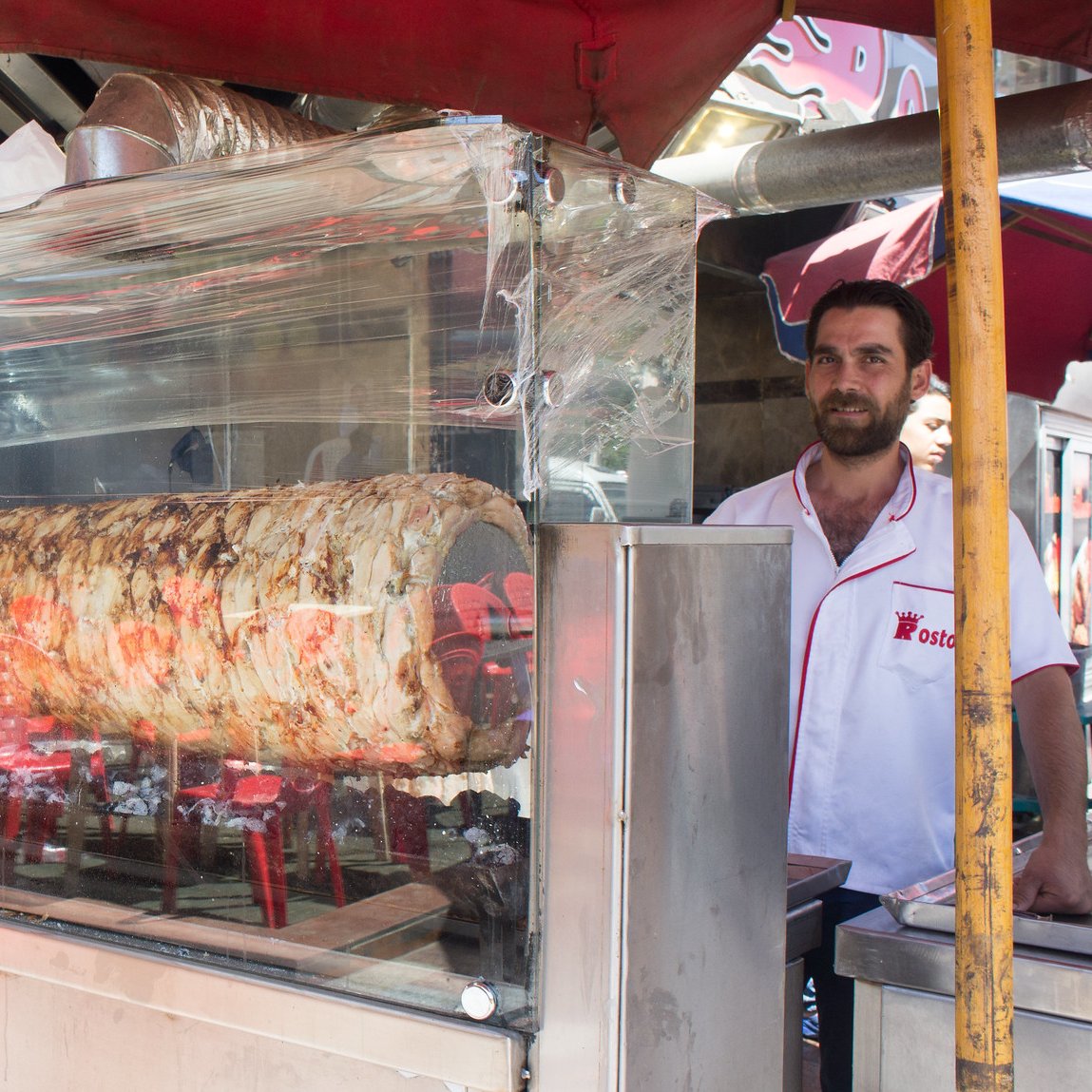 L'image montre un stand de nourriture de rue où un homme se tient derrière une vitrine en plastique transparente. À l'intérieur de la vitrine, il y a un grand morceau de viande en train de rôtir sur des braises, probablement un kebab ou un gyros. Le stand est couvert de parasols colorés, et il y a des affiches annonçant les plats proposés. L'ambiance semble vivante, typique des marchés animés de rues.