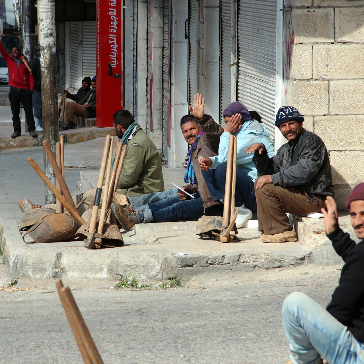الصورة تظهر مجموعة من الرجال يجلسون على الرصيف في منطقة حضرية. بعضهم يلوح بيده وكأنهم يتحدثون أو يتبادلون الحديث. يبدو أن لديهم أدوات عمل قربهم، مما يوحي بأنهم ربما كانوا ينتظرون فرصة للعمل. الشارع يبدو هادئًا ومظهراً لأبنية متوسطة الارتفاع. الأجواء تعكس نشاطًا ونمط حياة يومي في المدينة.