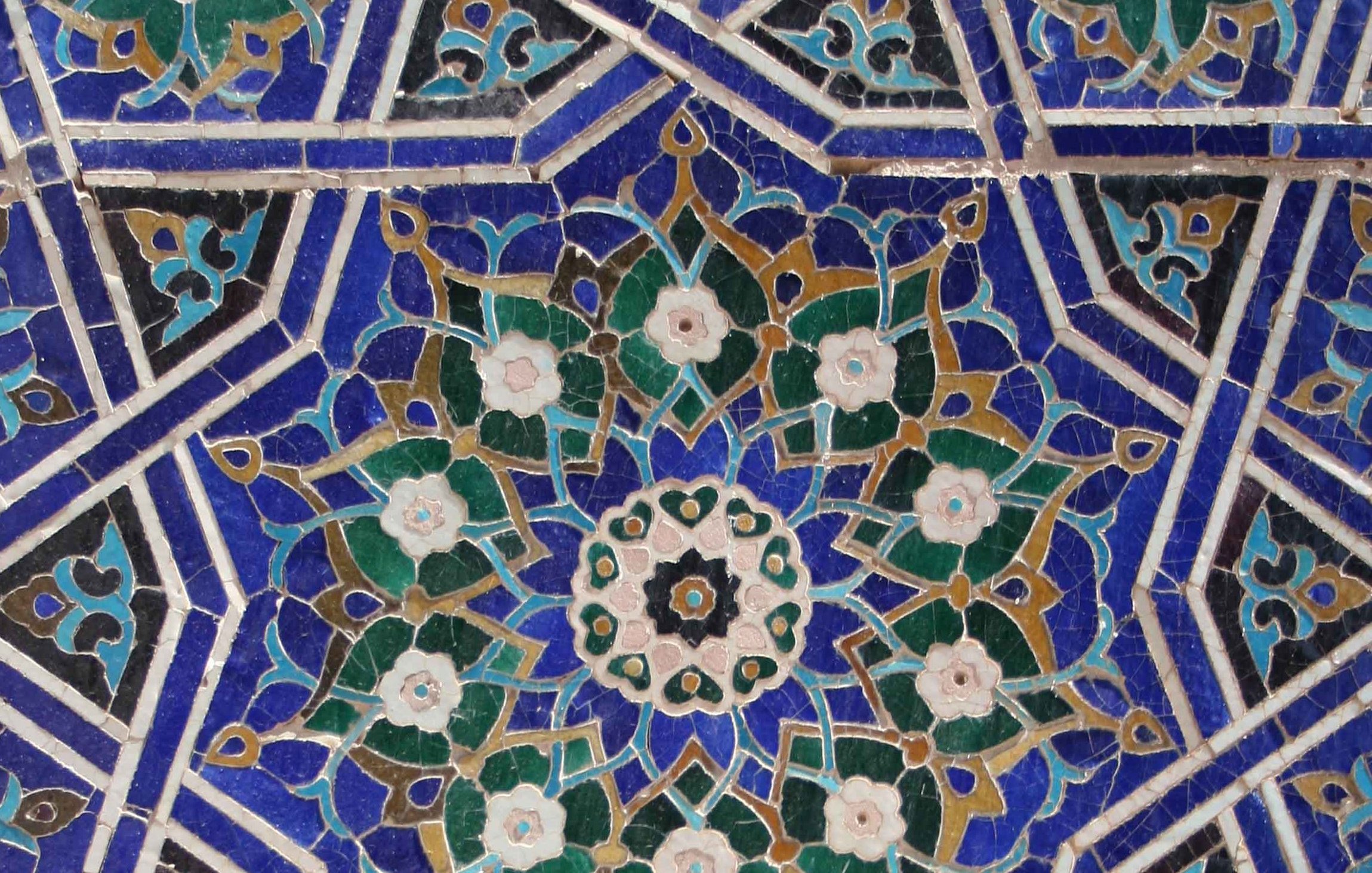 تظهر الصورة زخارف معمارية دقيقة، تتكون من أنماط هندسية وأزهار بألوان متنوعة مثل الأزرق والأخضر والكريمي. التصميم يبرز تداخل الأشكال بشكل متناظر، مع تفاصيل دقيقة تضيف جمالًا وتناسقًا للمظهر. يمكن رؤية تأثير الفنون الإسلامية التقليدية في هذا العمل، مما يعكس مهارة الحرفيين ودقتهم في تنفيذ مثل هذه الزخارف.