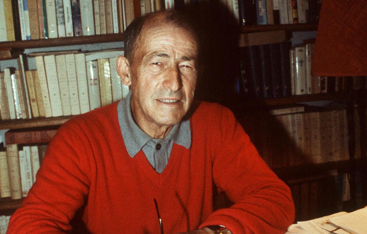 L'image montre un homme assis à un bureau, souriant, en train d'écrire sur une feuille de papier avec un stylo. Il porte un pull rouge et une chemise à col. En arrière-plan, on peut voir une bibliothèque remplie de livres. Sur le bureau, il y a également des feuilles de papier, peut-être des notes ou un manuscrit en cours de rédaction. L'éclairage crée une ambiance chaleureuse et studieuse.