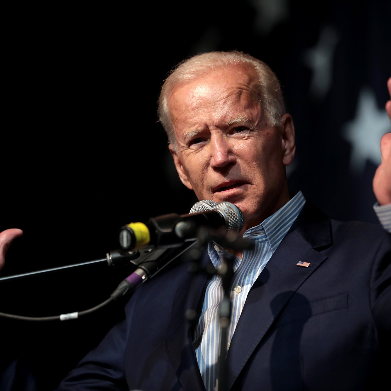 L'image montre un homme en train de donner un discours. Il est debout derrière un microphone et utilise ses mains pour gesticuler, ce qui suggère qu'il est passionné par ce qu'il dit. Il porte un costume bleu sombre avec une chemise à rayures. En arrière-plan, il y a un drapeau comportant des étoiles, ce qui pourrait donner un indice sur son contexte politique. L'atmosphère semble dynamique et engageante.