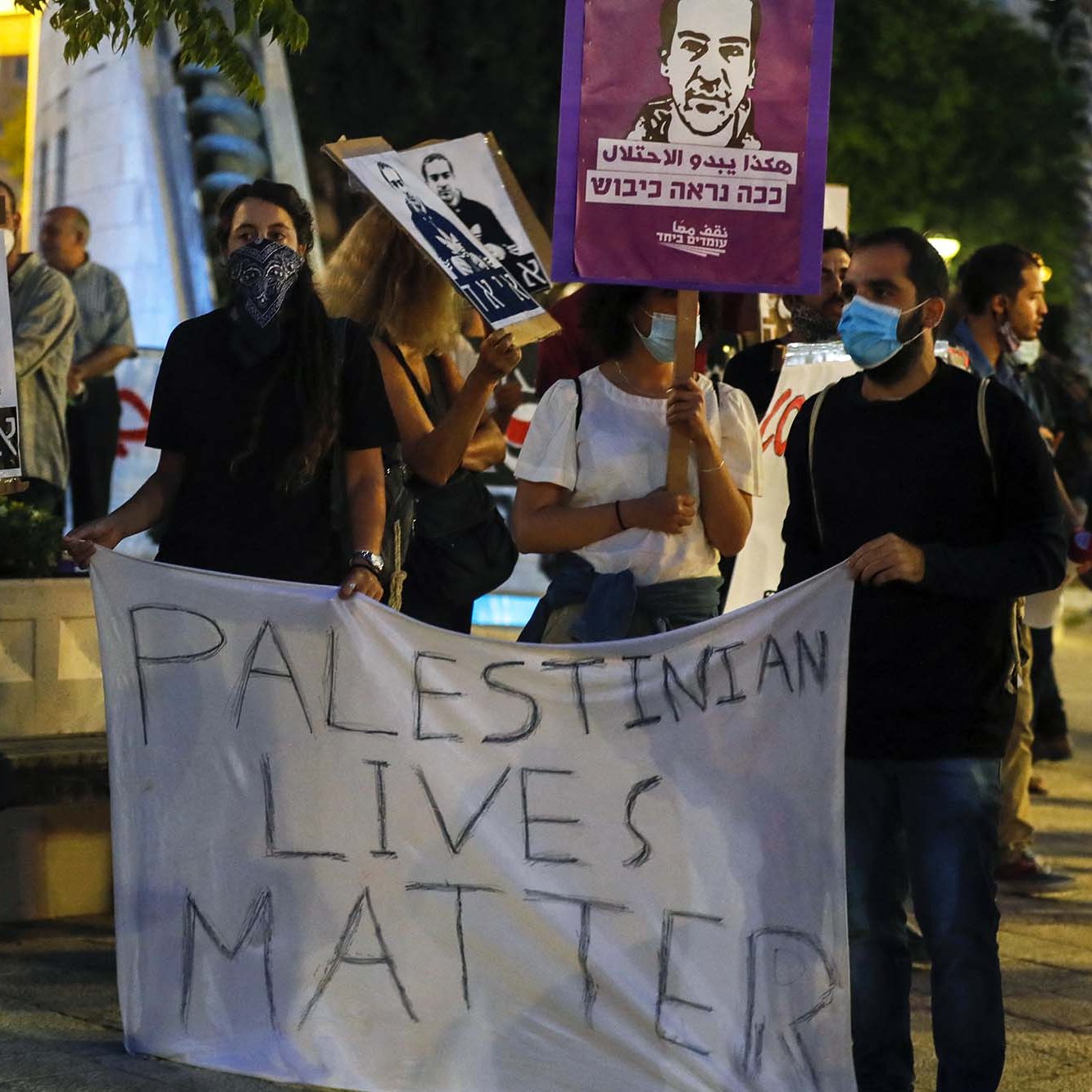 L'image montre un groupe de personnes manifestant. Elles tiennent des panneaux et une grande banderole sur laquelle est écrit "PALESTINIAN LIVES MATTER". Les participants portent des masques, et certains affichent des portraits d'individus. L'ambiance semble être celle d'une manifestation pour les droits des Palestiniens. Le décor urbain, avec des arbres et des lumières en arrière-plan, ajoute à l'atmosphère de l'événement.
