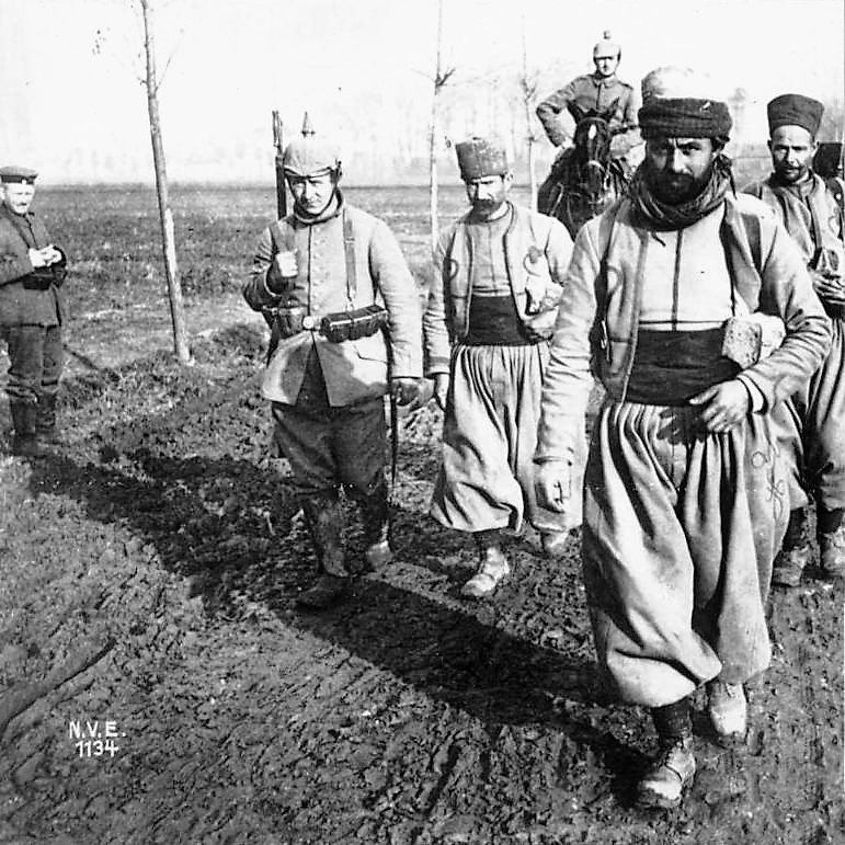 L'image montre un groupe de soldats marchant sur un terrain boueux. Ils portent des uniformes traditionnels, comprenant des pantalons larges et des bonnets. L'un des soldats est monté sur un cheval, tandis qu'un autre se tient en retrait, à l'écart du groupe. Le décor semble rural, avec quelques arbres en arrière-plan, suggérant un contexte militaire historique. L'atmosphère est sérieuse et solennelle, reflétant les conditions de la guerre.