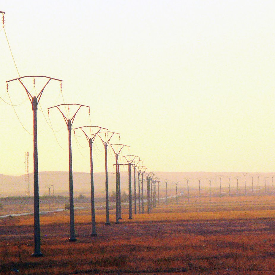 L'image montre une perspective sur un paysage de campagne, dominé par des pylônes électriques s'étendant sur une longue distance. Le sol est principalement sec et herbeux, avec des teintes dorées, suggérant la lumière douce d'un lever ou d'un coucher de soleil. En arrière-plan, on peut apercevoir des collines ou des montagnes floues, contribuant à une ambiance calme et désertique. L'ensemble évoque un sentiment d'immensité et de tranquillité.