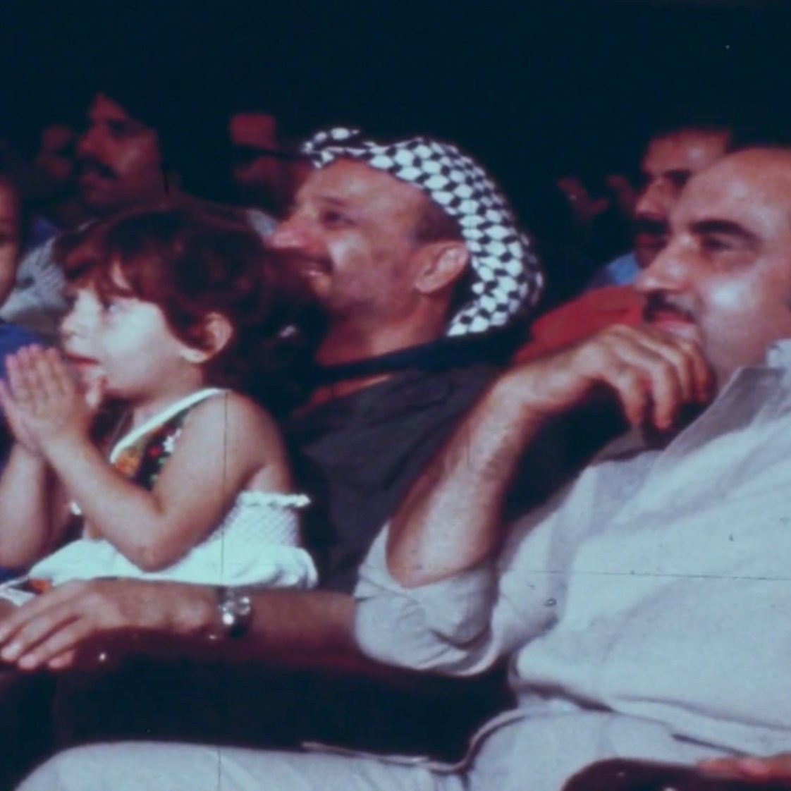L'image montre un groupe de personnes assises dans une salle, probablement lors d'un événement. Au premier plan, deux enfants, un garçon et une fille, semblent applaudir. Le garçon porte un maillot de bain et la fille est habillée avec une petite robe. Derrière eux, un homme portant un keffieh (le couvre-chef traditionnel) sourit. À sa droite, un autre homme est assis, regardant également vers le devant. L'ambiance paraît festive et conviviale.