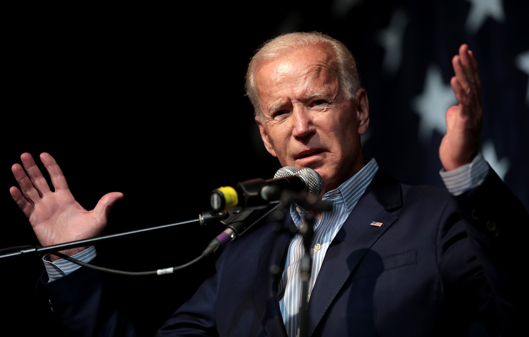 L'image montre un homme en train de donner un discours. Il est debout derrière un microphone et utilise ses mains pour gesticuler, ce qui suggère qu'il est passionné par ce qu'il dit. Il porte un costume bleu sombre avec une chemise à rayures. En arrière-plan, il y a un drapeau comportant des étoiles, ce qui pourrait donner un indice sur son contexte politique. L'atmosphère semble dynamique et engageante.