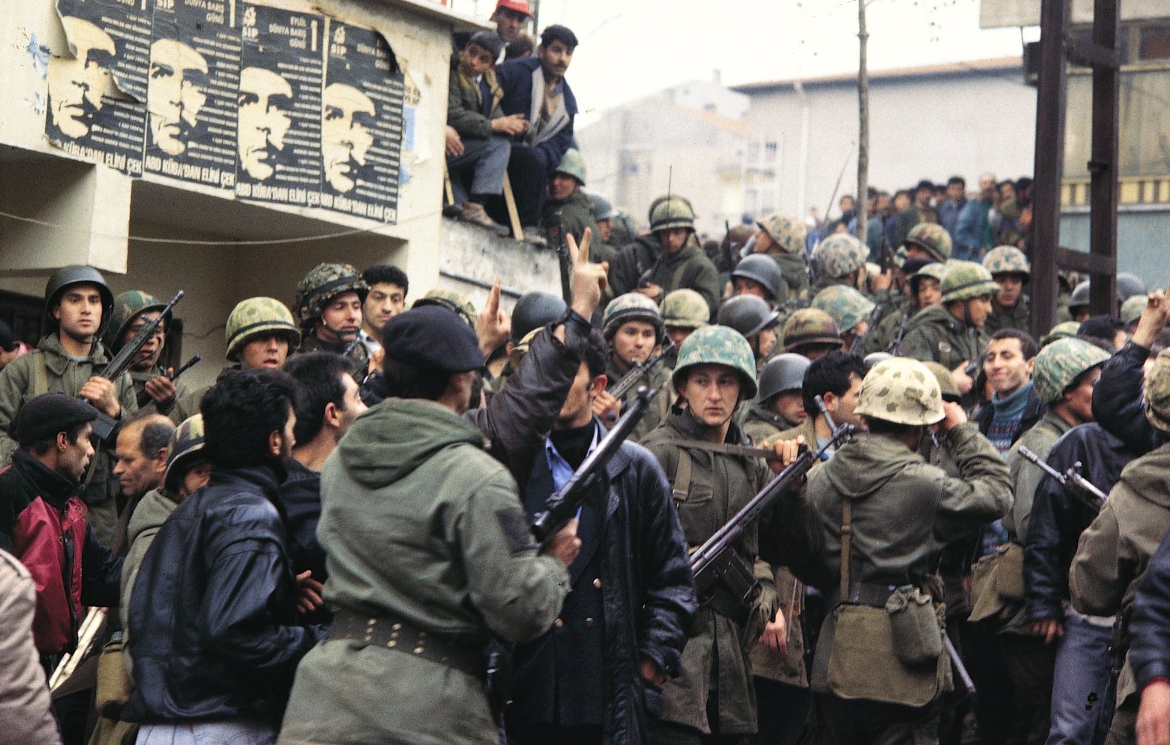 L'image montre un groupe de soldats en uniforme, portant des casques et armés, réunis autour d'une foule. On peut voir des hommes en civil, certains semblent s'opposer aux militaires. En arrière-plan, des affiches sont visibles sur le mur, peut-être représentant des figures politiques ou des messages de protestation. L'atmosphère est tendue et indique un moment de conflit ou de manifestation. Les visages des individus expriment des émotions variées, allant de la détermination à l'inquiétude.