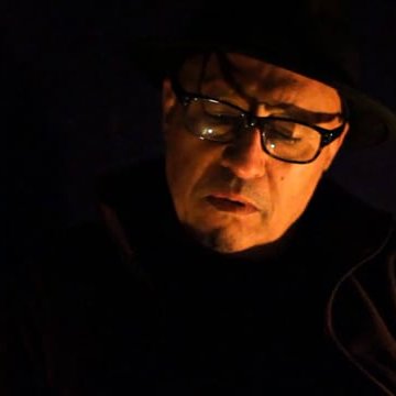 L'image montre un homme avec des lunettes et un chapeau. Il semble pensif et est plongé dans une lumière tamisée, ce qui crée une atmosphère sombre et introspective. Son expression faciale suggère qu'il est en train de réfléchir profondément ou de vivre un moment d'émotion.