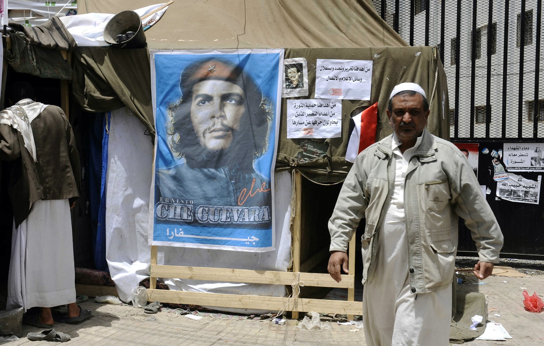 L'image montre un campement avec une tente, devant laquelle se trouve une affiche représentant un homme avec un béret, souvent associé à Che Guevara. À côté de l'affiche, on peut voir des affiches supplémentaires et une personne marchant, vêtue d'une tenue clair. L'ambiance semble refléter un contexte politique ou social, probablement avec des éléments de protestation ou de revendication.