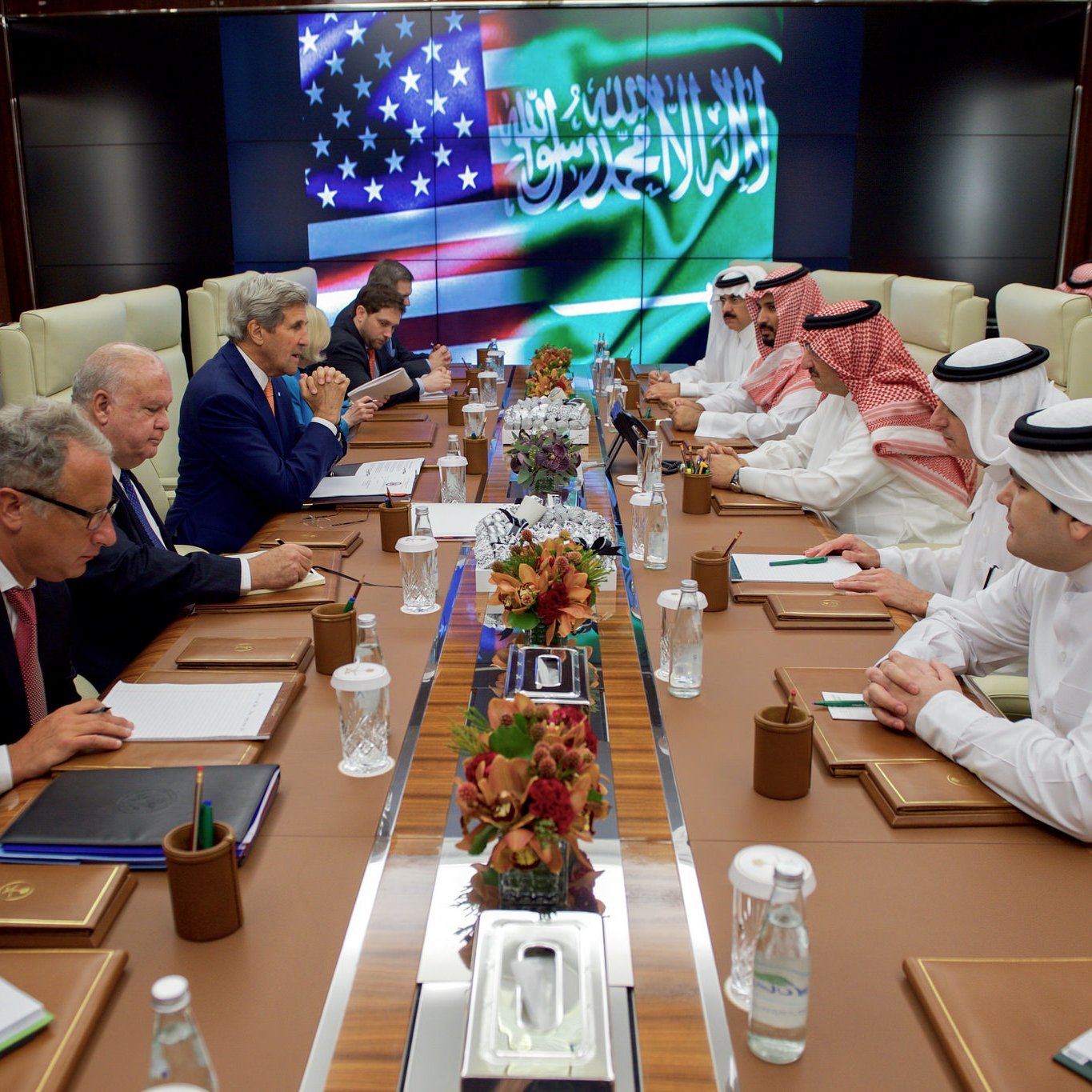 L'image montre une réunion diplomatique dans une salle de conférence. On peut voir plusieurs personnes assises autour d'une longue table. À l'arrière-plan, il y a un écran affichant les drapeaux des États-Unis et d'Arabie saoudite. Les participants semblent être engagés dans une discussion sérieuse, avec des documents et des ordinateurs portables devant eux. L'atmosphère est formelle et professionnelle. Les membres de la réunion sont habillés en costumes et en vêtements traditionnels arabes.