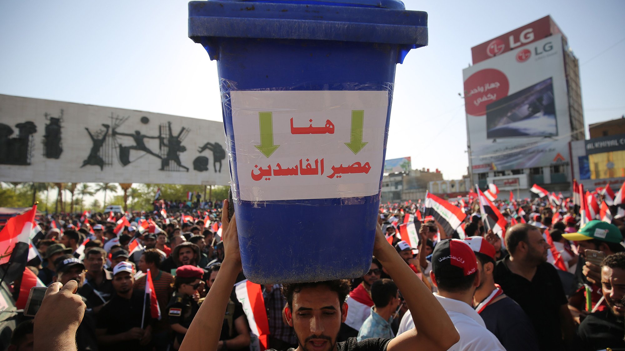 L'image montre une grande manifestation avec une foule rassemblée. Au premier plan, un homme porte un grand seau bleu sur sa tête, qui affiche des inscriptions en arabe. Derrière lui, on peut voir de nombreux drapeaux, probablement ceux de l'Égypte, et des affiches graphiques sur un mur. La scène dégage une atmosphère de protestation, avec des gens mobilisés pour une cause.