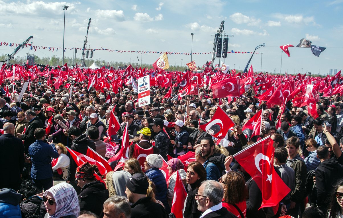 The image shows a large crowd gathered in an outdoor setting, with many people holding Turkish flags. The atmosphere appears to be lively and energetic, with individuals visibly engaged in the event. Some are holding banners, while others are raising their flags high. The background features clear skies and some structures, possibly related to the event or gathering. Overall, it depicts a significant public demonstration or celebration, likely related to a national or cultural event in Turkey.