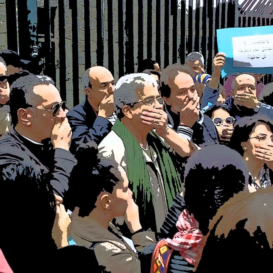 L'image montre un groupe de personnes réunies dans un cadre extérieur. Beaucoup d'entre elles se tiennent avec la main sur la bouche, semblant exprimer un silence ou protester. En arrière-plan, on aperçoit un panneau qui semble contenir un message. L'ambiance générale semble sérieuse et engagée, suggérant une forme de mobilisation ou de manifestation. Les participants semblent concentrés et solidaires.