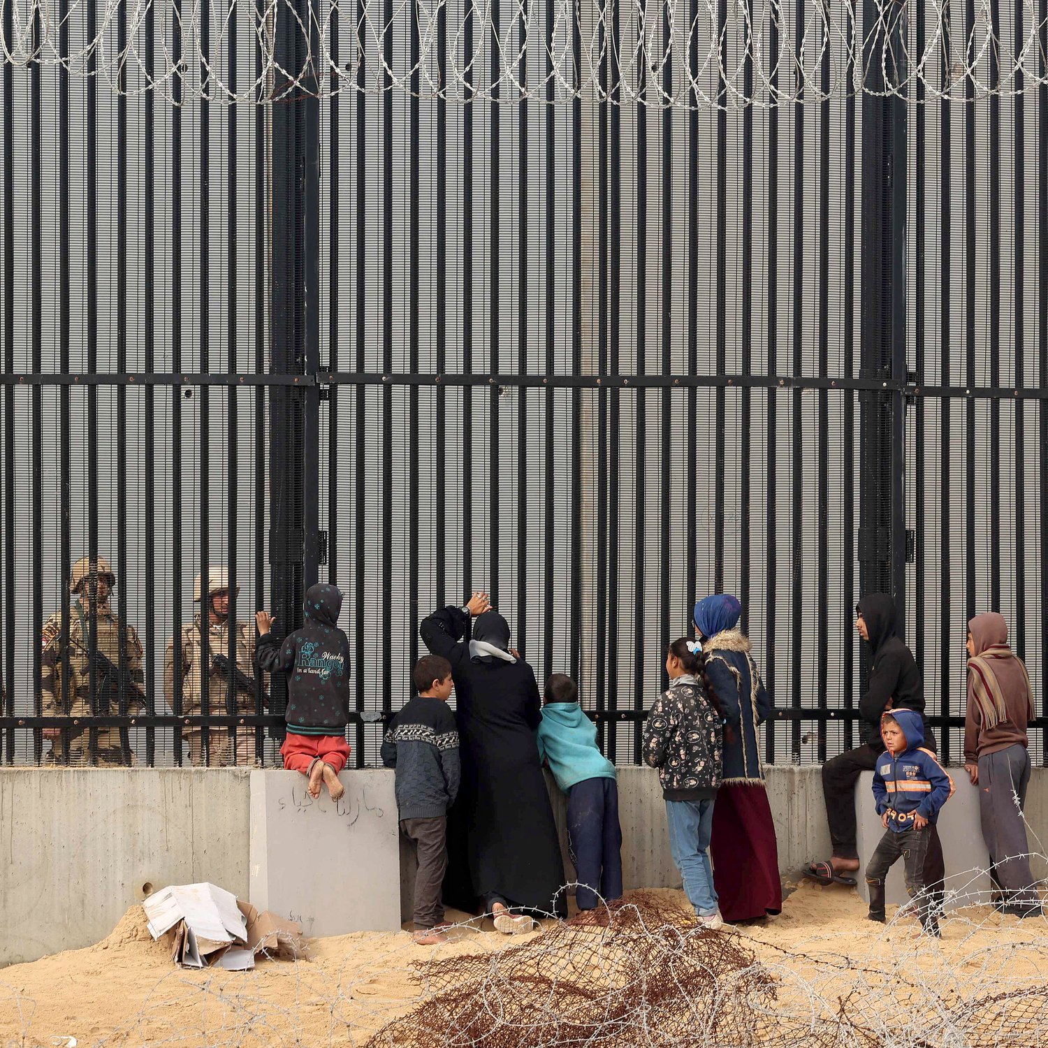 The image depicts a group of people, including women and children, standing in front of a tall, barred fence. On the other side of the fence are soldiers or guards, who appear to be watching the group. The scene conveys a sense of separation and tension, with barbed wire in the foreground emphasizing the barriers between the two sides. The individuals on the left seem to be engaging with the guards, possibly seeking assistance or communication. The overall atmosphere suggests a complex and challenging situation, likely related to migration or security issues.