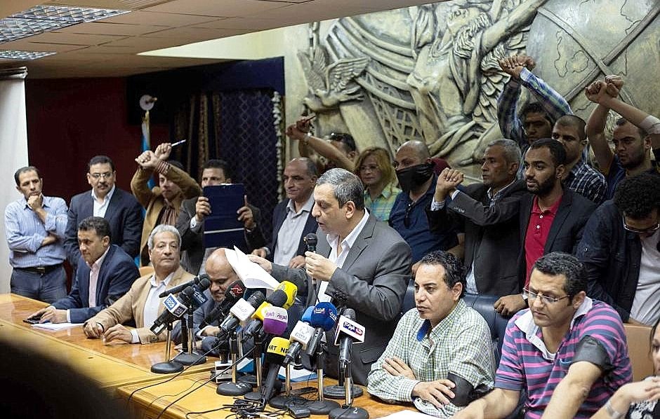 L'image montre une conférence de presse ou une réunion. Un homme se tient au centre, parlant devant un groupe de journalistes, avec plusieurs microphones devant lui. Derrière lui, des personnes semblent exprimer leur soutien ou leur désaccord, levant les poings. L'arrière-plan présente une sculpture en relief qui ajoute de la profondeur à la scène. L'atmosphère semble tendue et engagée.
