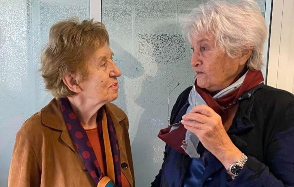 L'image montre deux femmes âgées engagées dans une conversation. Elles semblent amicales et souriantes. L'une porte une veste marron avec un foulard à pois, tandis que l'autre est habillée de manière similaire, avec une écharpe. L'arrière-plan est flou, mettant en valeur leur interaction.