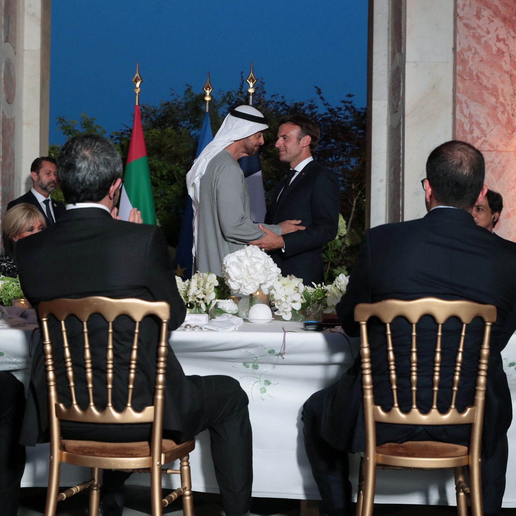 L'image montre une scène formelle dans un cadre élégant, probablement lors d'un dîner ou d'une réception. Au centre, deux hommes se serrent la main. On peut voir des drapeaux en arrière-plan, suggérant un contexte diplomatique. À la table, plusieurs personnes assistent à l'événement, avec une attention particulière sur les deux protagonistes. L'ambiance est chaleureuse et conviviale, marquée par des éléments de décoration soignés, comme des fleurs.
