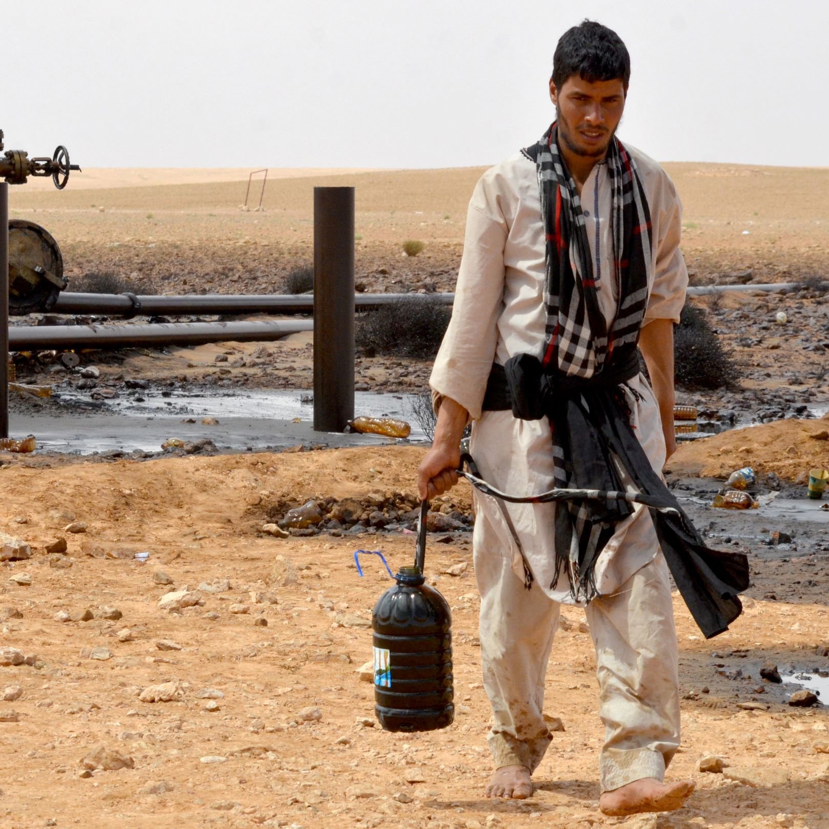 L'image montre un homme marchant à travers un paysage désertique. Il porte des vêtements traditionnels et a un seau noir dans une main. En arrière-plan, on peut voir des installations liées à l'industrie pétrolière, avec des tuyaux et des structures. Le sol est parsemé de pierres et il y a des traces de pétrole sur le terrain. L'environnement semble aride et désertique.