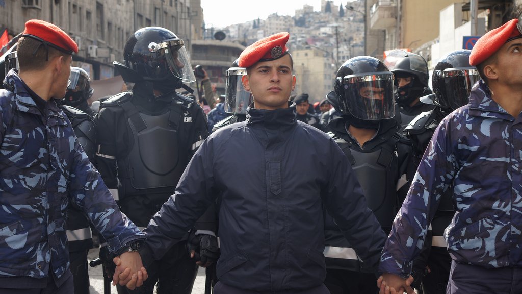 L'image montre un groupe de policiers ou de soldats, avec des uniformes distinctifs, se tenant les mains. Au centre, un homme en majorette avec un béret se distingue. Derrière eux, des membres des forces de l'ordre portent des casques et des équipements de protection. L'environnement semble urbain, avec des bâtiments en arrière-plan. L'atmosphère peut suggérer une situation de tension ou de manifestation.