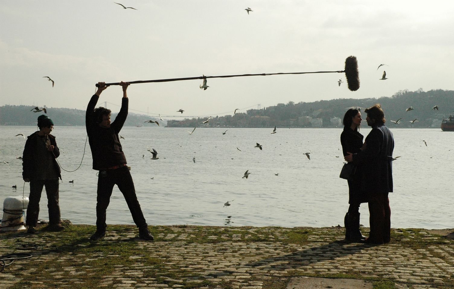L'image montre une scène de tournage près de l'eau. On peut voir un homme tenant un perche avec un micro au-dessus de deux personnes qui semblent discuter. Ces personnes sont au premier plan, tandis qu'à l'arrière-plan, on aperçoit des oiseaux volant autour et un paysage flou. Un autre homme, au loin, semble superviser le tournage. Le temps paraît légèrement nuageux, ajoutant une atmosphère particulière à la scène.
