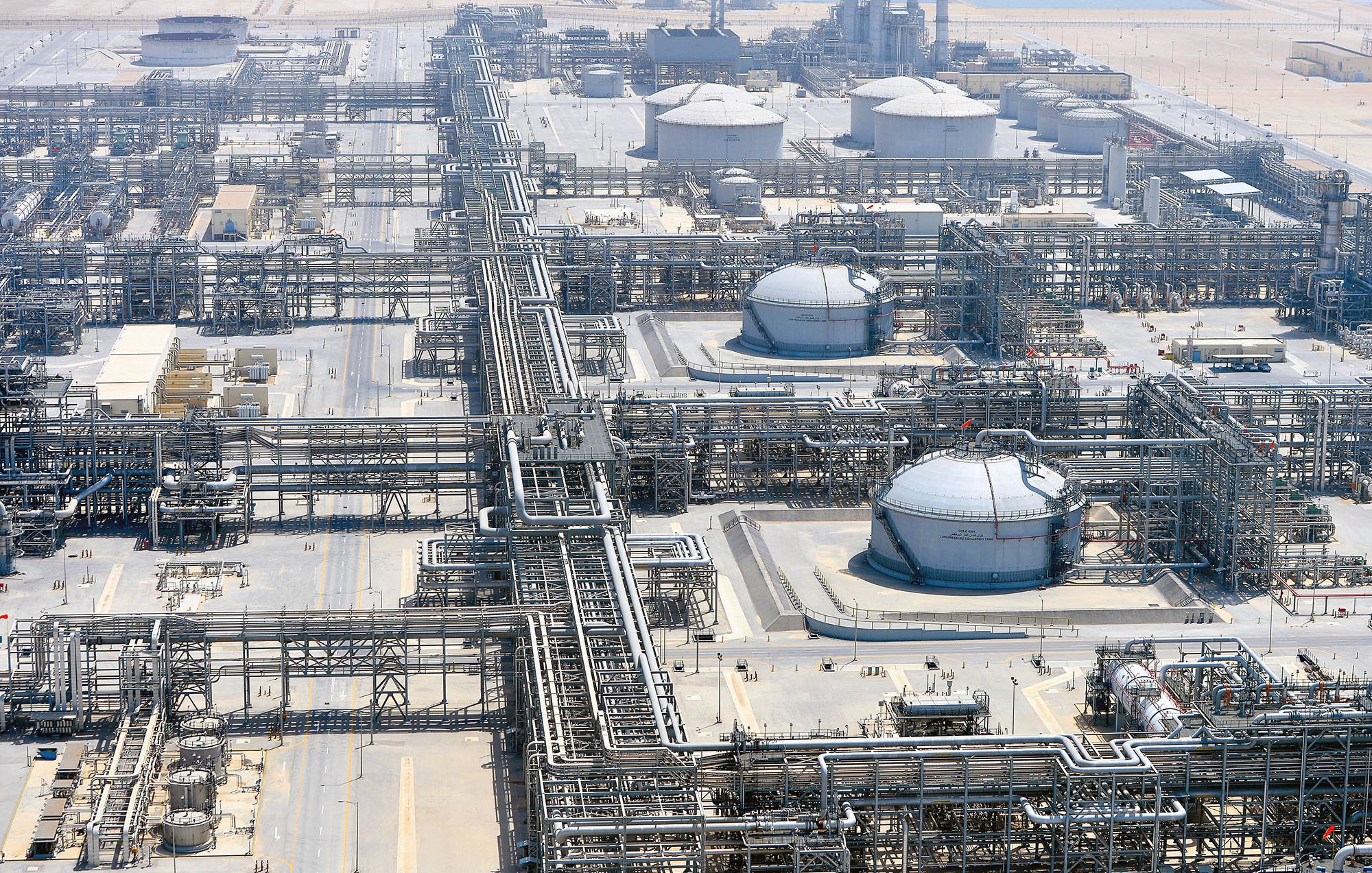 الصورة تُظهر منظرًا جويًا لمصنع صناعي ضخم، يبدو أنه يتعلق بقطاع النفط أو الغاز. يمكن رؤية مجموعة من الخزانات الدائرية الكبيرة، بالإضافة إلى شبكة معقدة من الأنابيب والبنى التحتية الأخرى. المنطقة محاطة بأرض صحراوية، مما يدل على أن المصنع يقع في منطقة جافة. يركز التصميم على الكفاءة في نقل وإدارة المواد الخام، مما يشير إلى مستوى عالٍ من التكنولوجيا والهندسة.