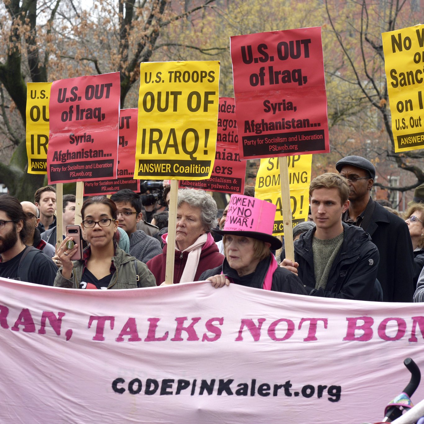 L'image montre une manifestation où des personnes tiennent des pancartes avec des slogans anti-guerre. Plusieurs affiches demandent le retrait des troupes des États-Unis d'Irak, de Syrie et d'Afghanistan. Les manifestants semblent déterminés et unis dans leur message, qui appelle à la paix, avec un grand banner rose affichant "IRAN, TALKS NOT BOMBS" et le site web de CODEPINK. L'atmosphère semble à la fois sérieuse et engagée.