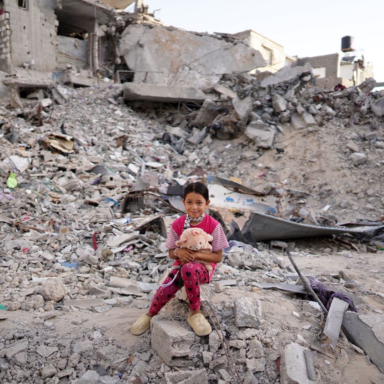L'image montre une jeune fille assise sur des décombres, entourée de ruines. Elle porte des vêtements colorés et tient un jouet, semblant à la fois vulnérable et résiliente au milieu du chaos. La scène évoque des sentiments de tristesse et de perte, en raison de la destruction visible sur son environnement.
